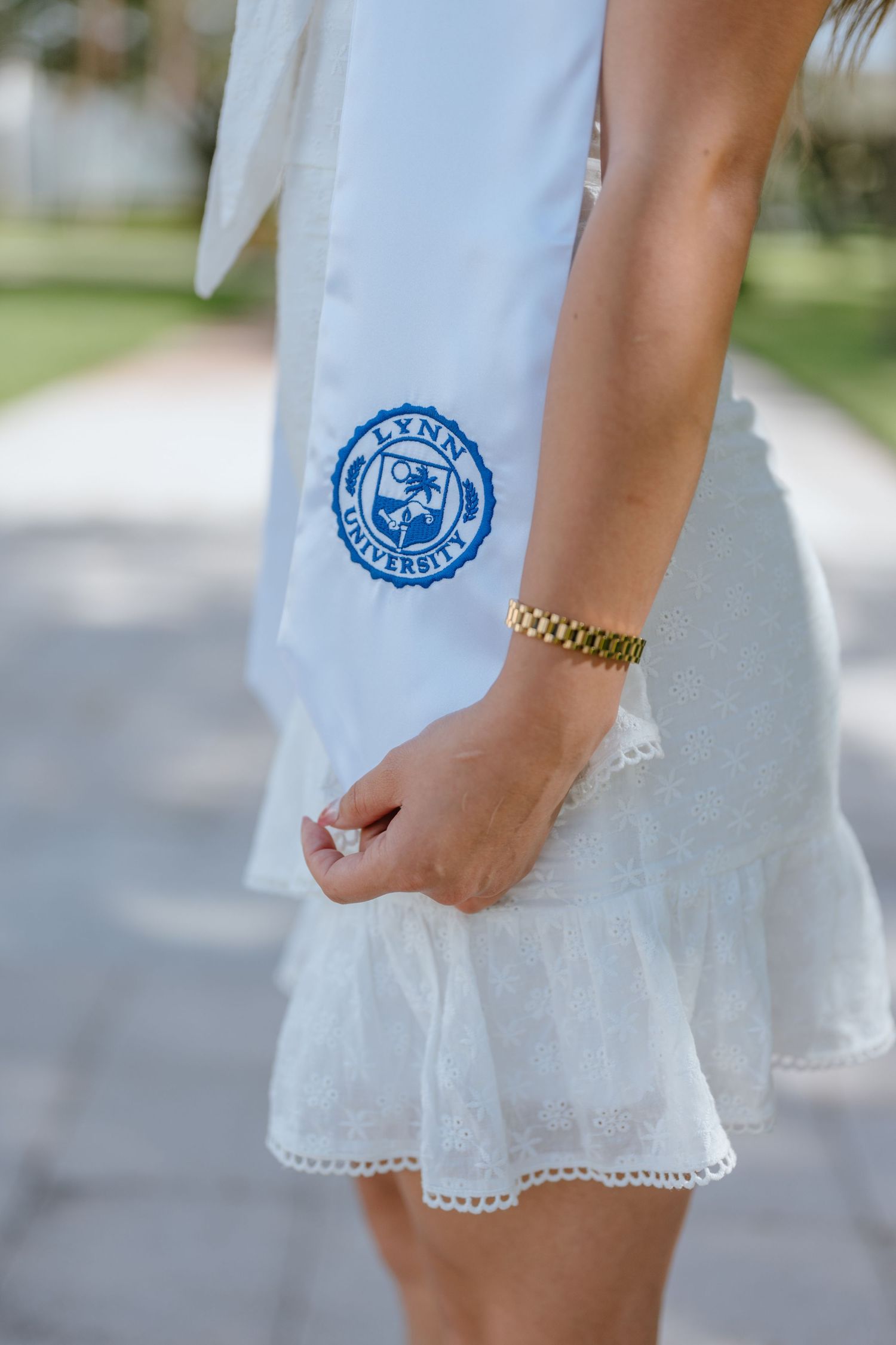 Close-up of Lynn University graduation stole with blue seal worn with white dress and gold bracelet.