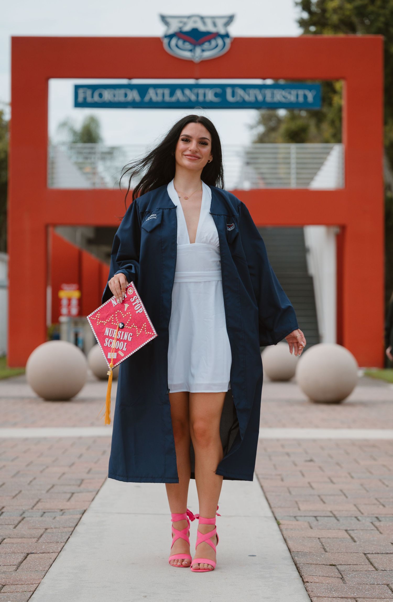 Graduate in navy gown holds nursing school cap while walking past Florida Atlantic University entrance sign.