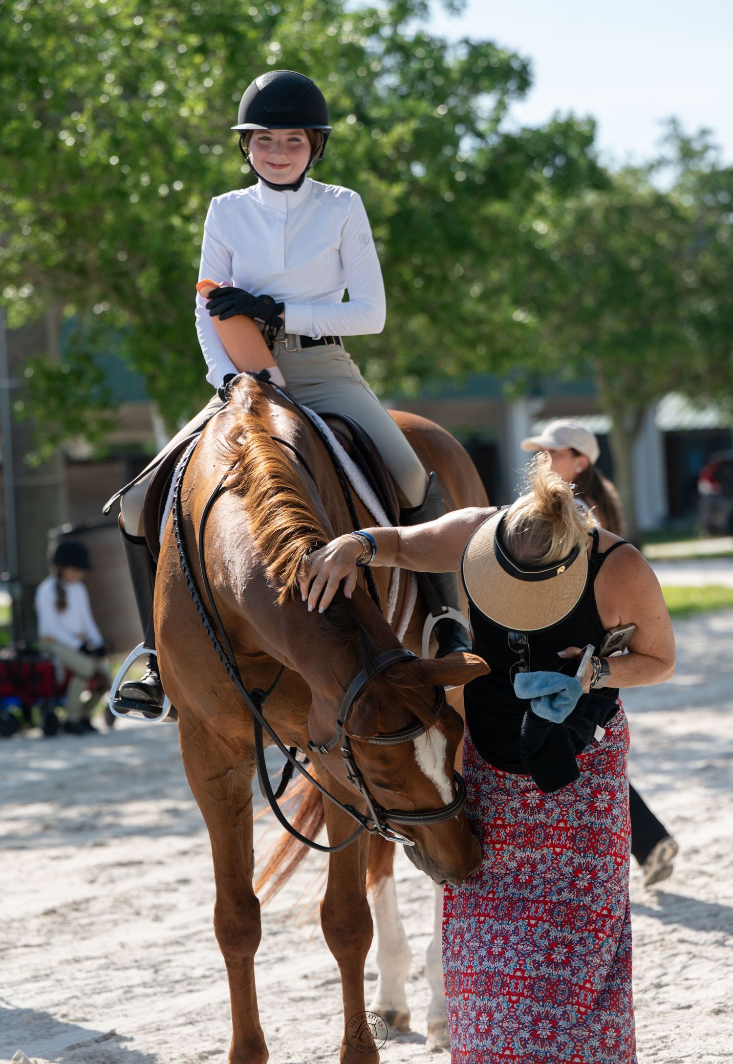 Smiling young female equestrian on chestnut horse receiving assistance from trainer at outdoor horse show.