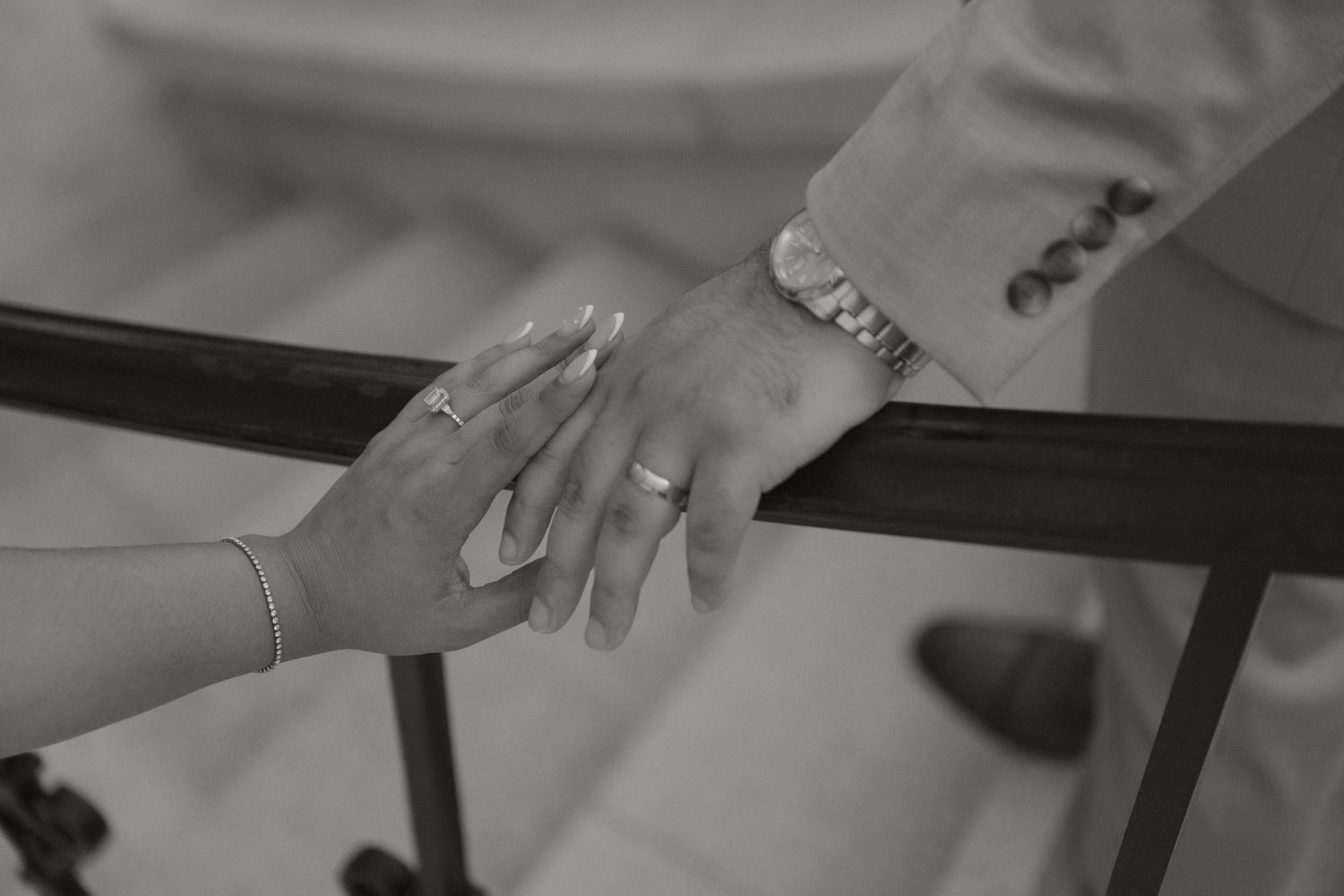 Black and white close-up of couple holding hands on staircase railing, showing engagement ring and bracelet.