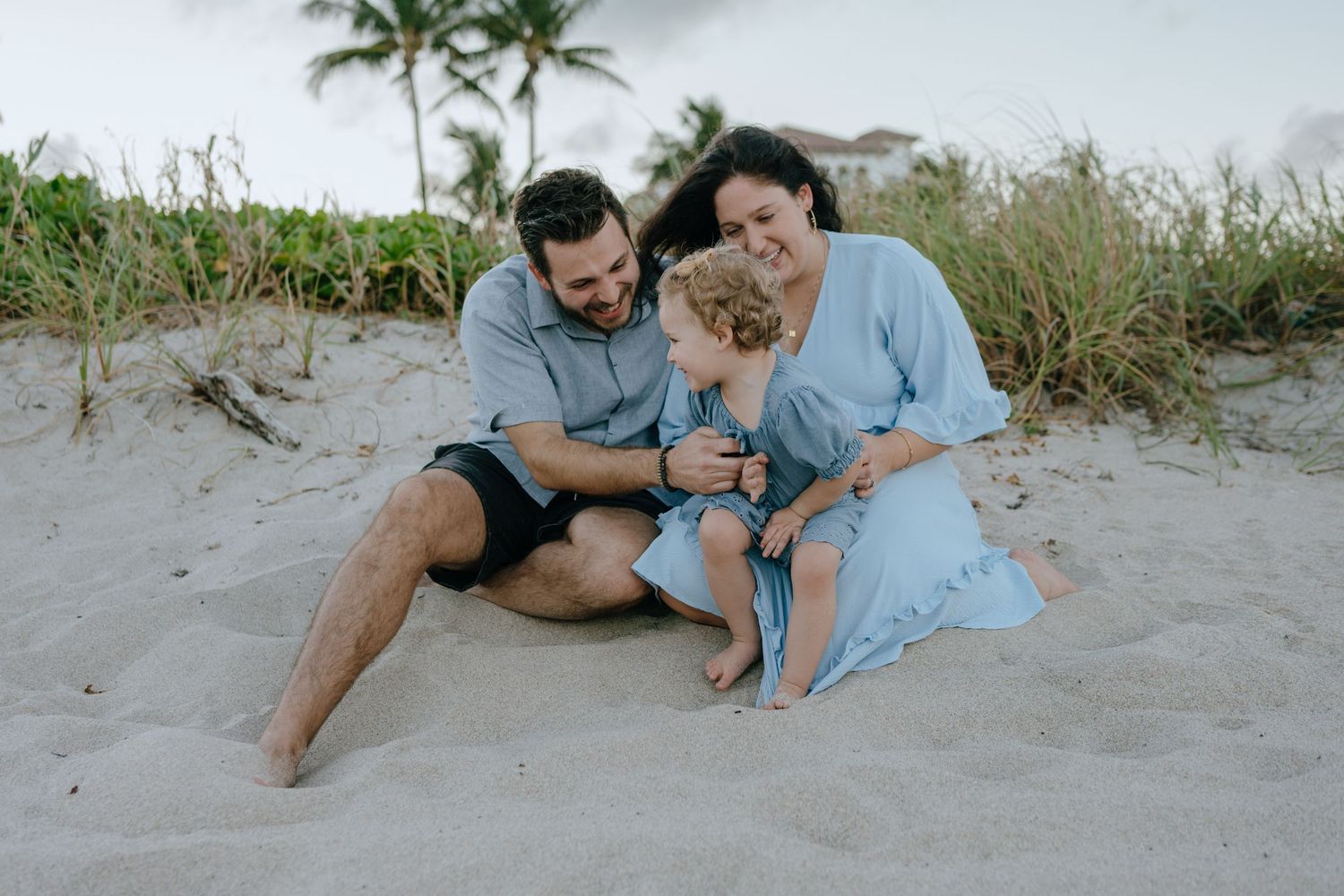 Young family of three sitting together on sandy beach dunes, sharing a joyful moment at twilight.