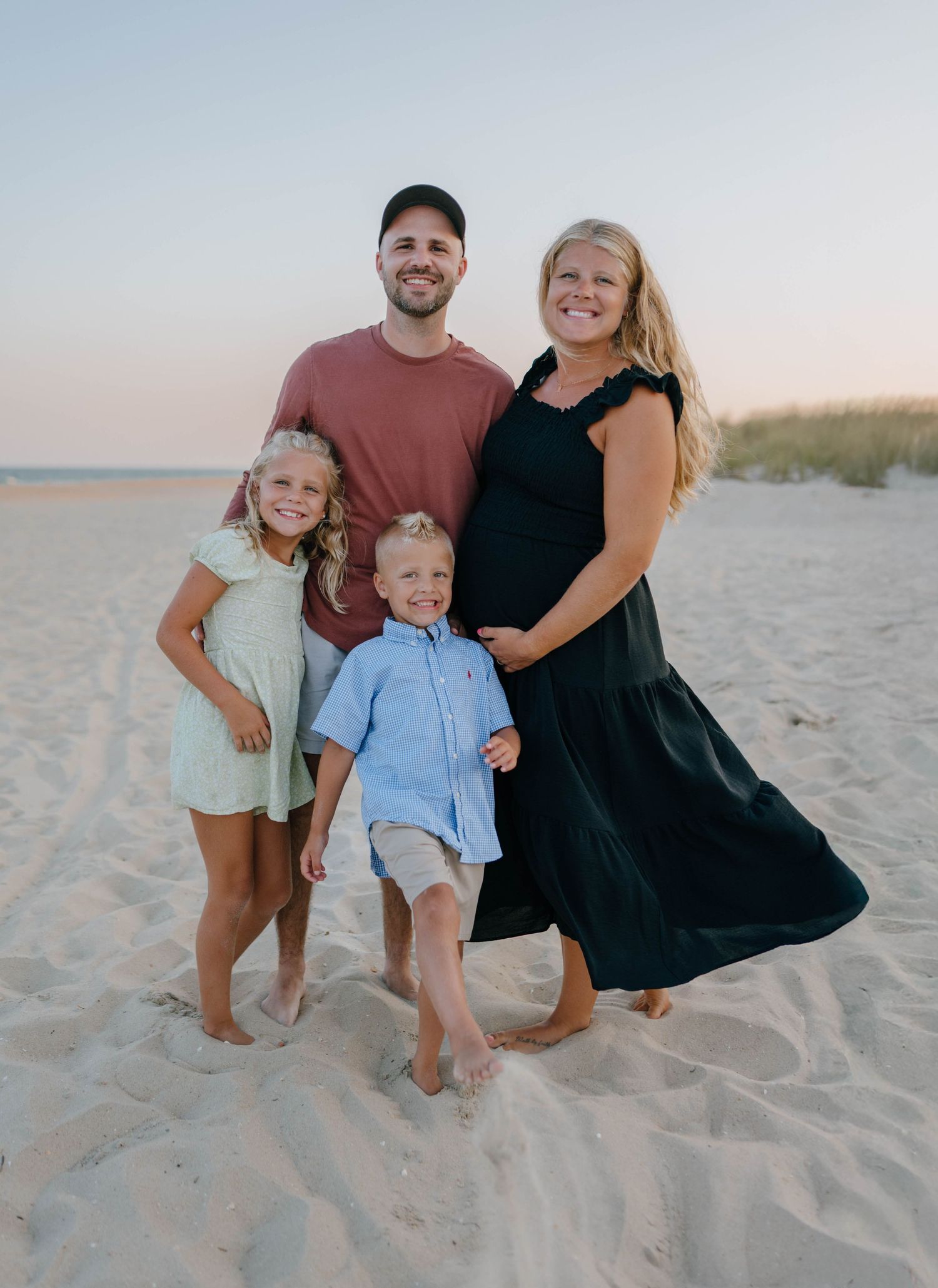Happy family of four poses on sandy beach at sunset, pregnant mother in black dress with husband and two young children.