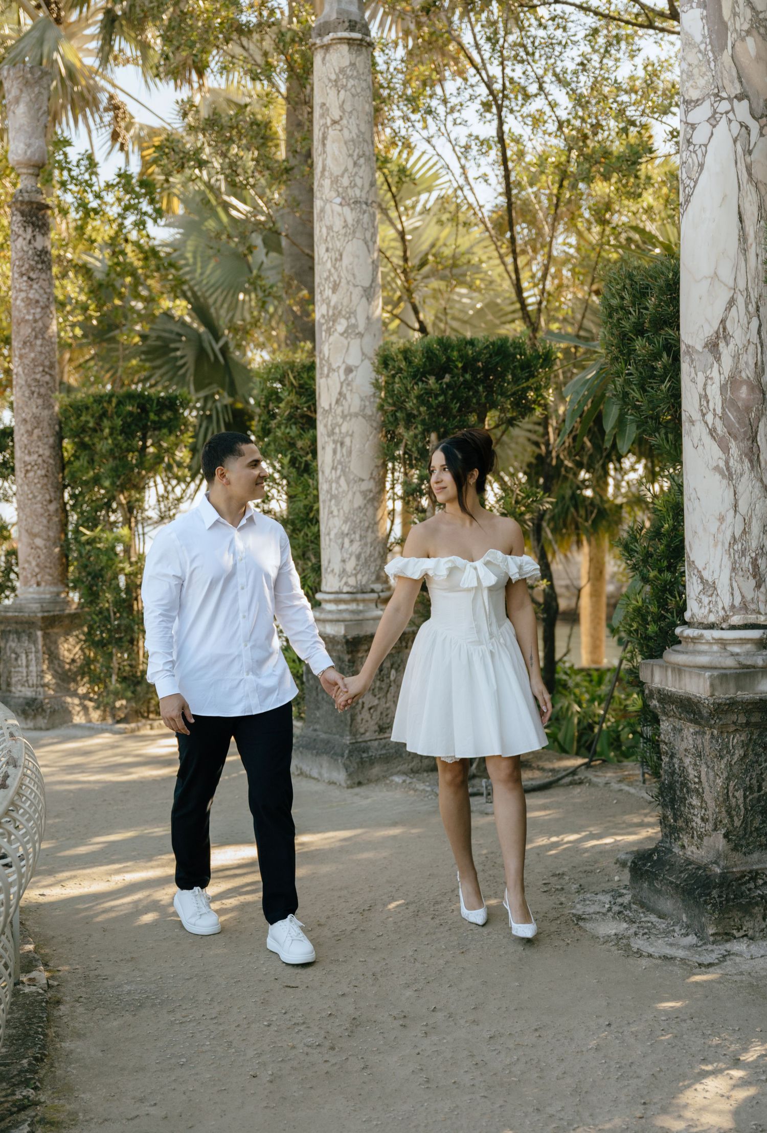Couple walking hand in hand through sunlit garden colonnade, woman in white off-shoulder mini dress.