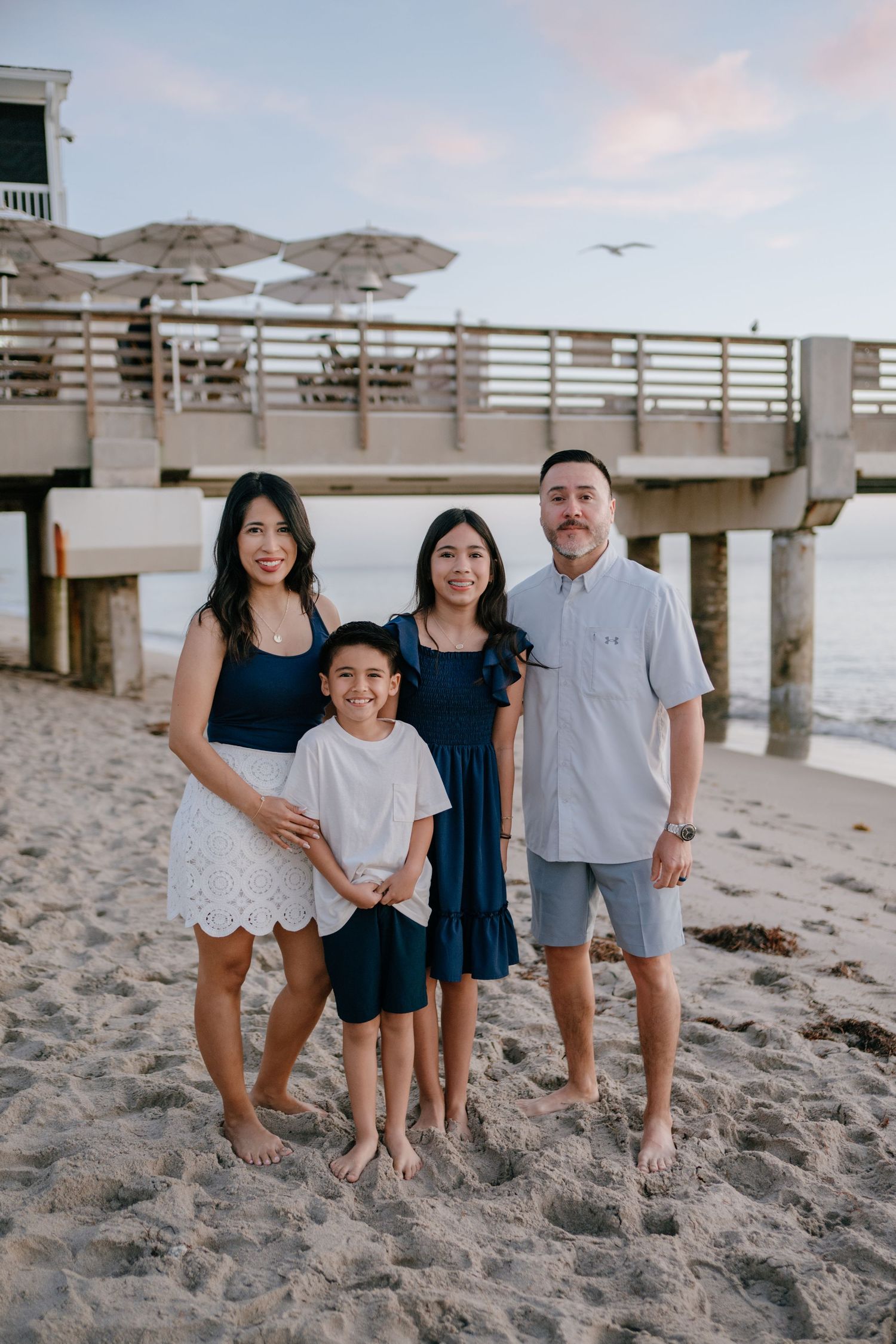 Family of four in navy and white outfits smiling together on sandy beach at sunset near pier.