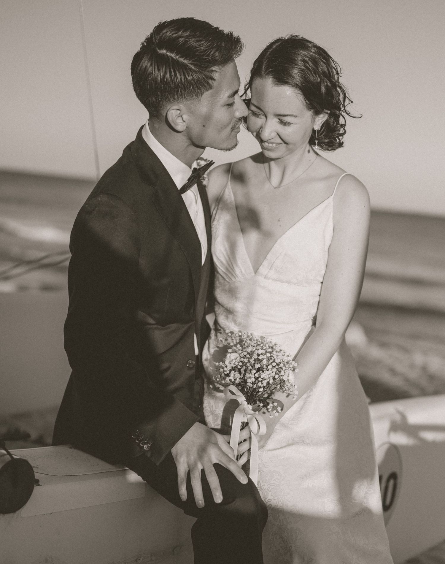 Black and white photo of newlywed couple sharing a romantic moment on a sailboat by the beach.