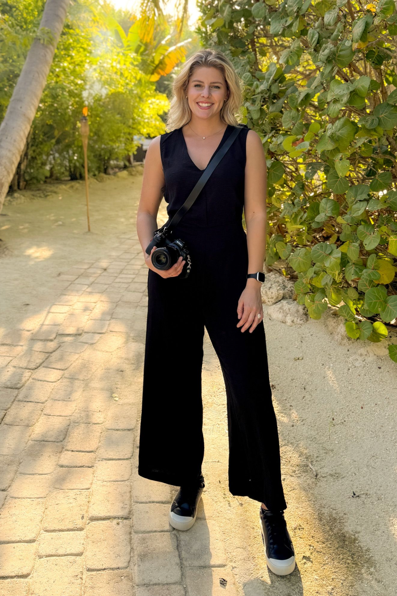 Blonde female photographer in a black jumpsuit holding a camera on a tropical sunlit pathway with lush greenery.