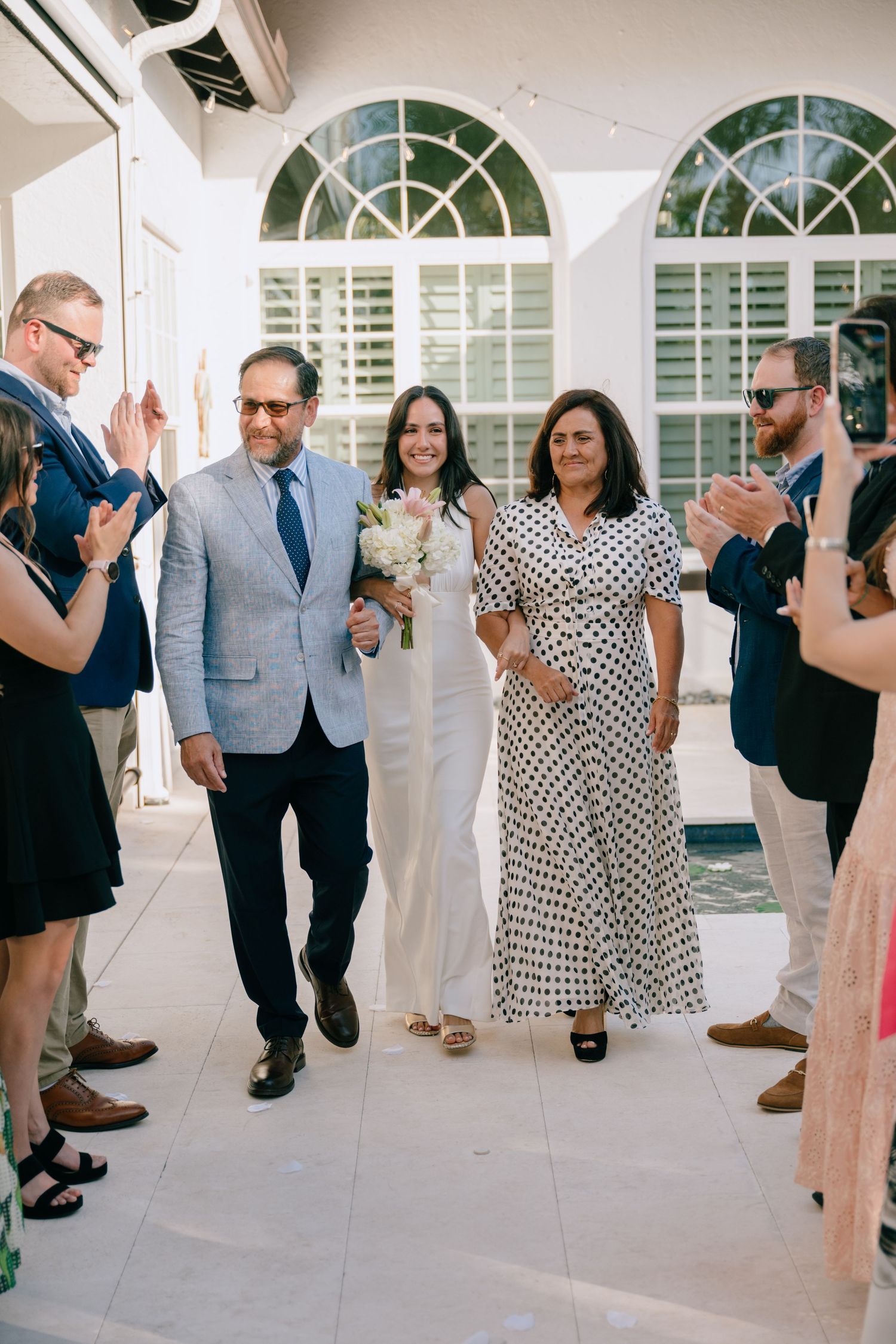 Bride walks down the aisle escorted by both parents at an elegant outdoor wedding ceremony.