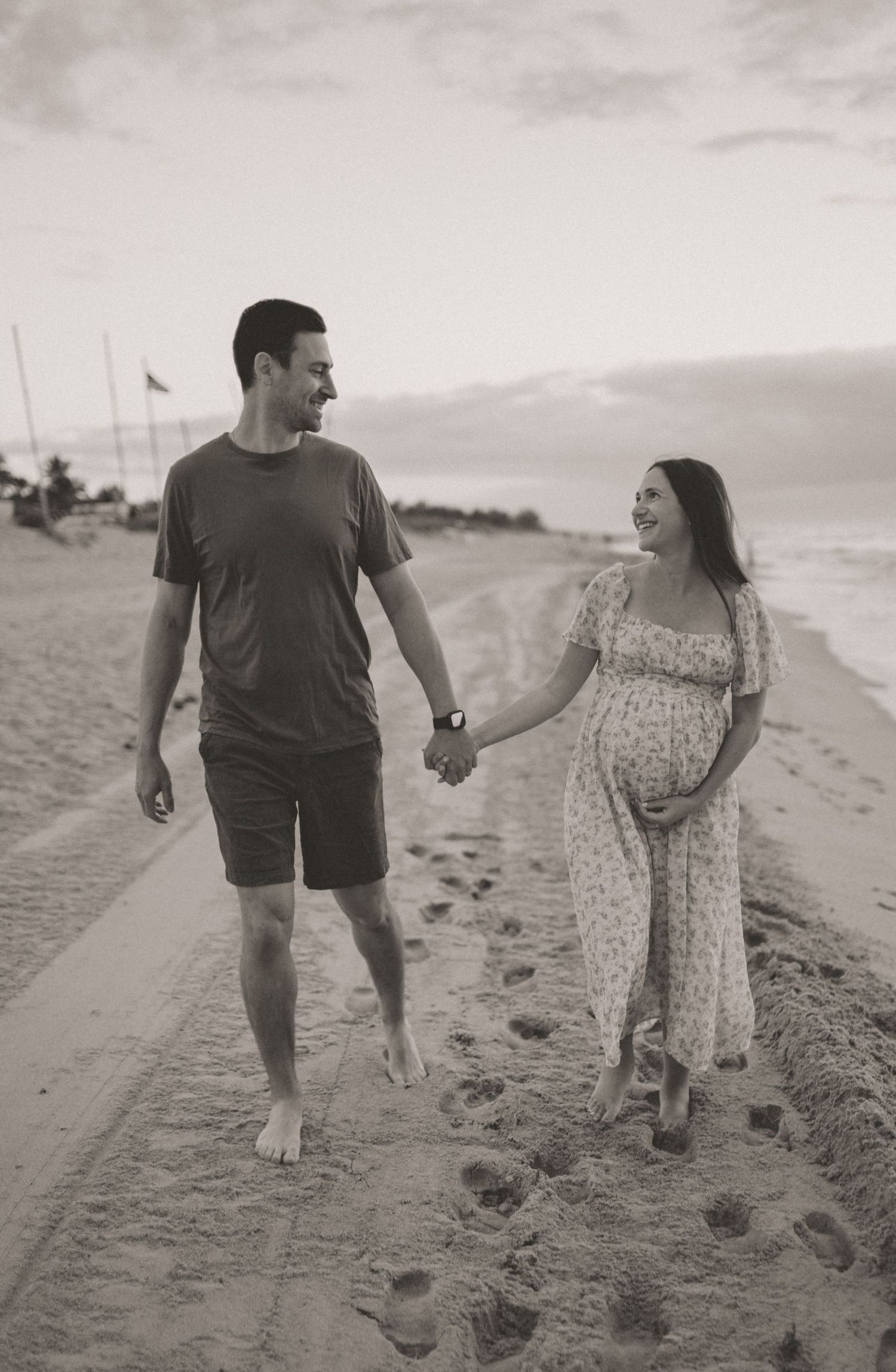 Black and white photo of smiling couple walking hand in hand on beach during maternity shoot.
