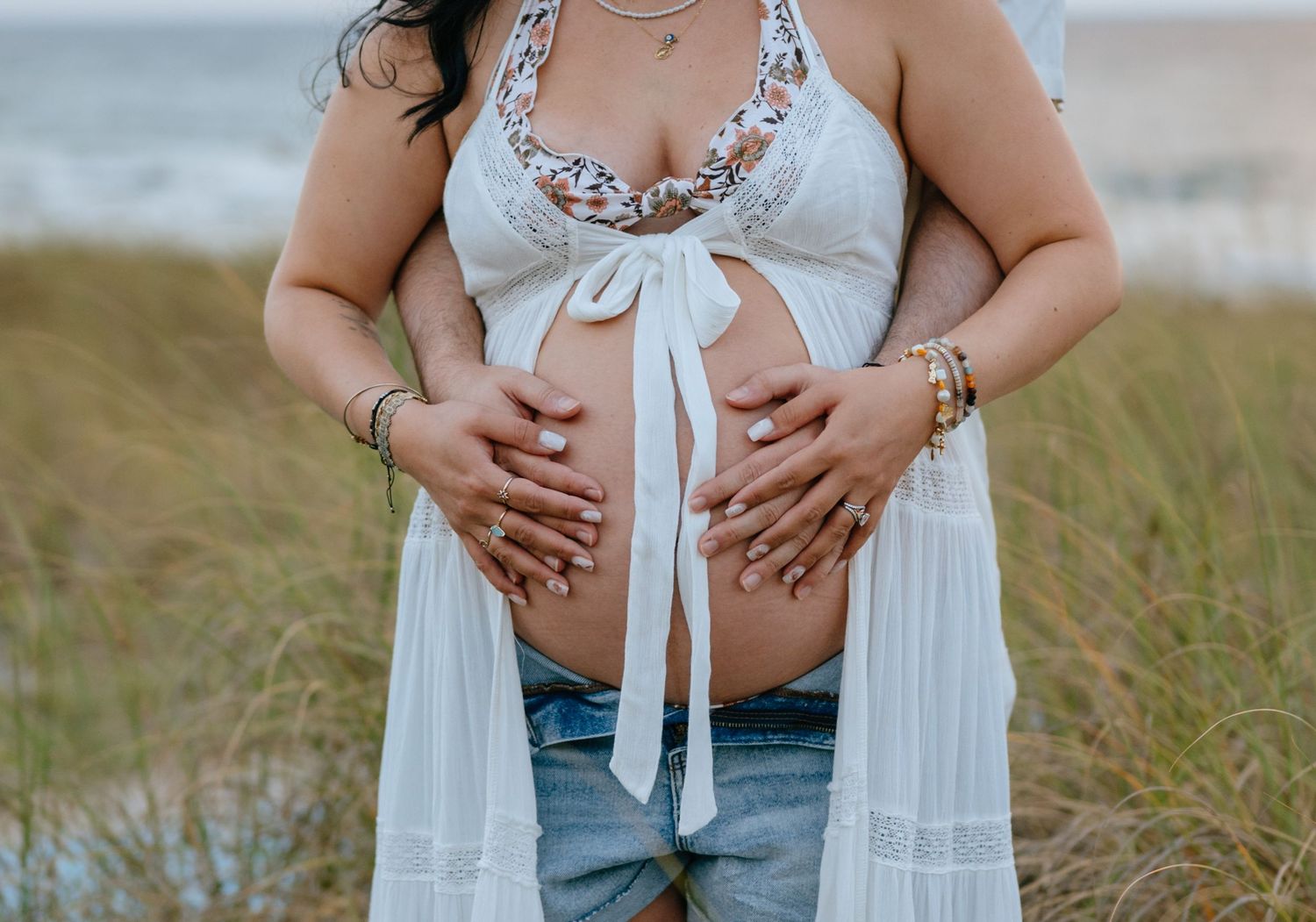Pregnant woman in white lace top and denim shorts holding bare baby bump outdoors.