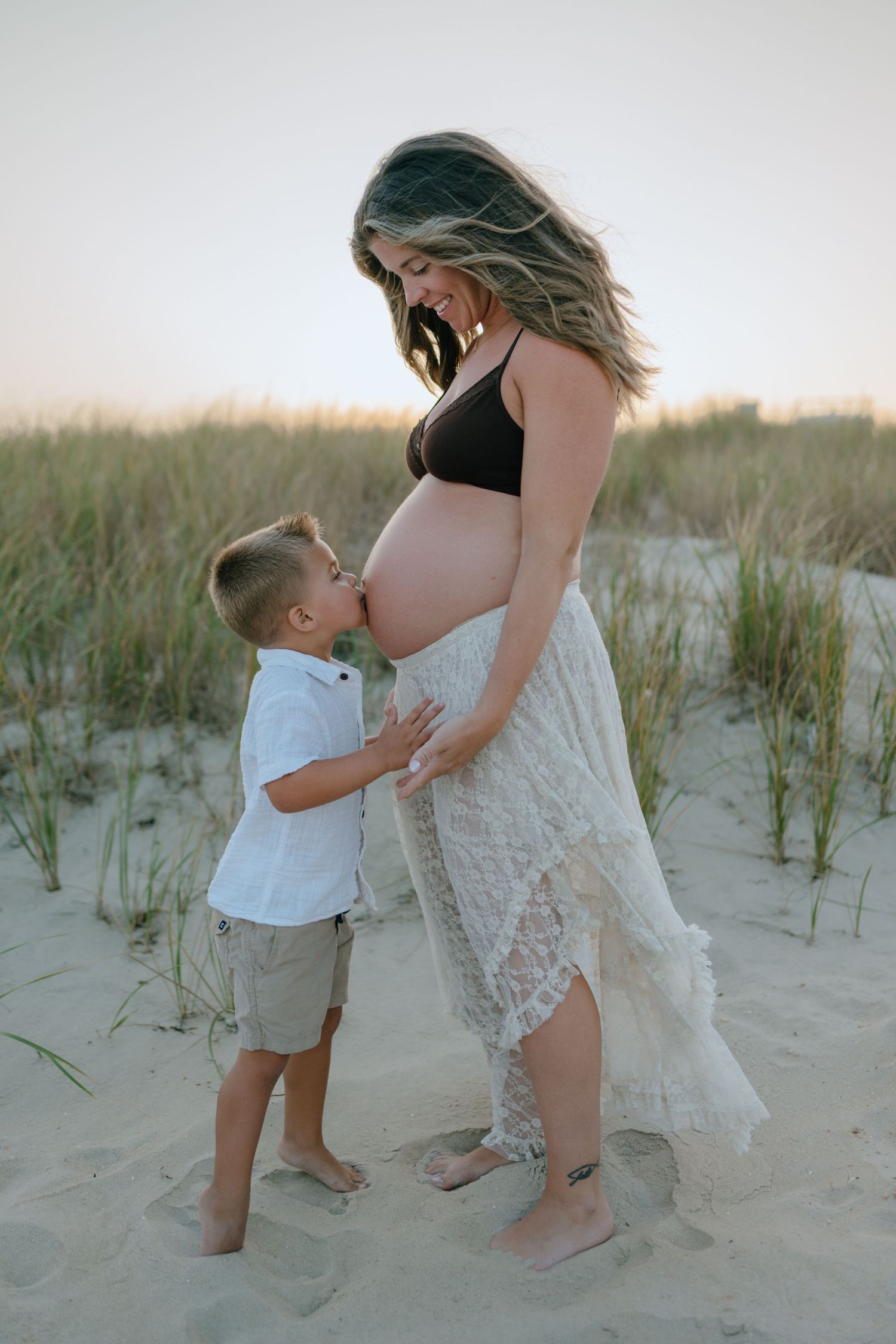 Young boy kisses pregnant mother's bare belly on sandy beach dunes at golden hour.