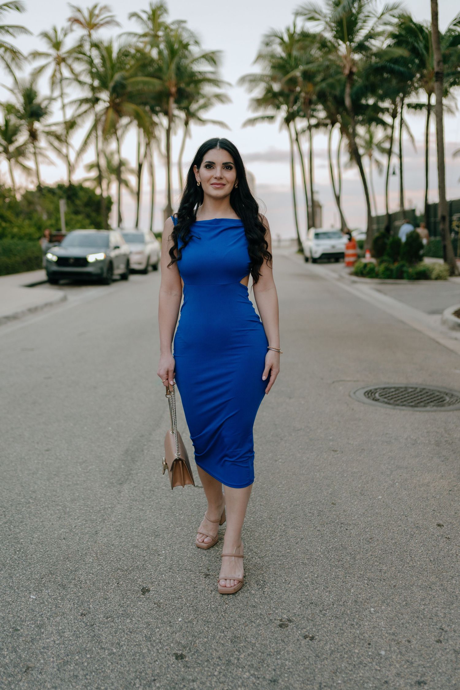 Woman in royal blue midi dress walking palm-lined street holding woven bag.