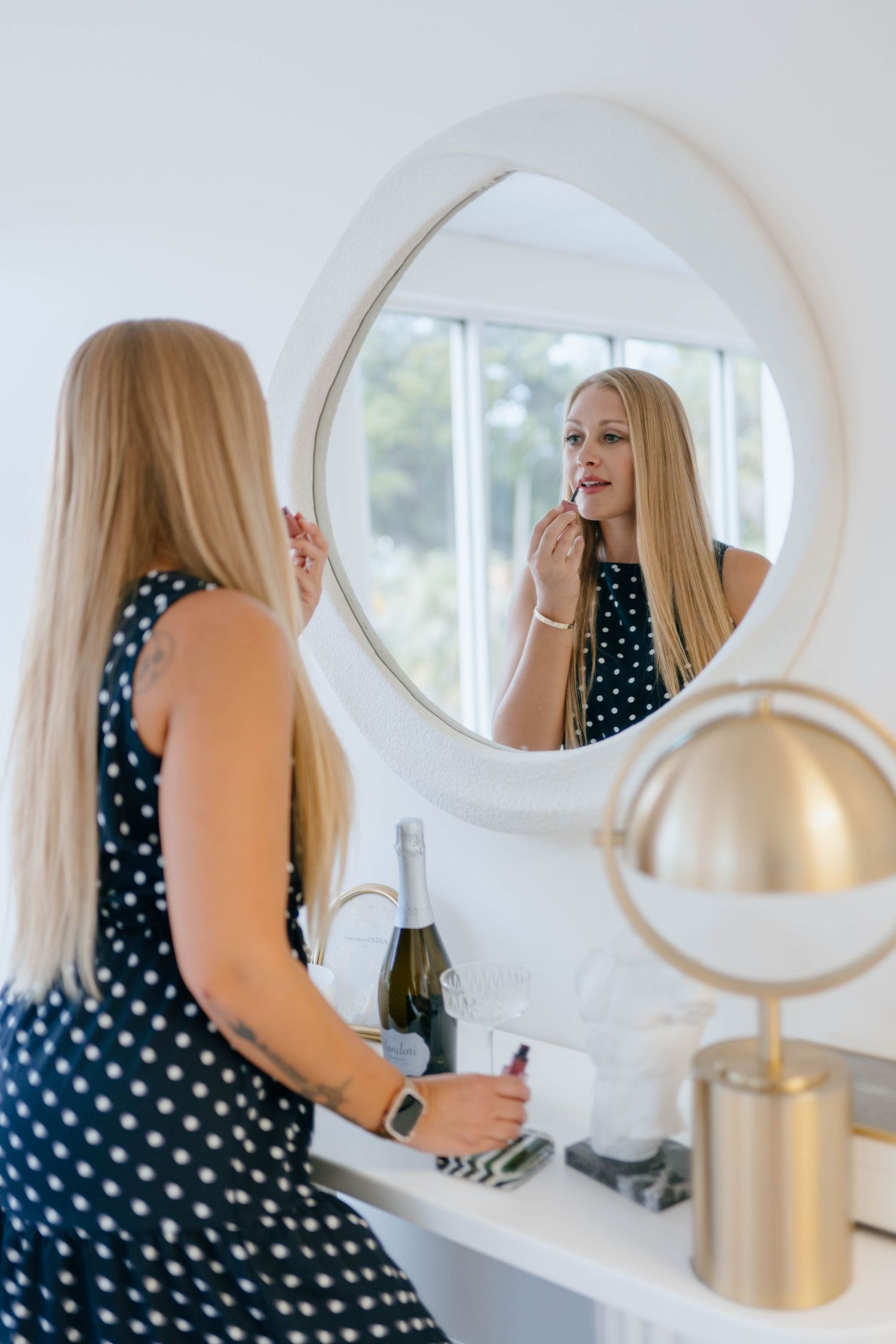 Blonde woman applying lipstick while looking into a round white mirror at a vanity with champagne nearby.
