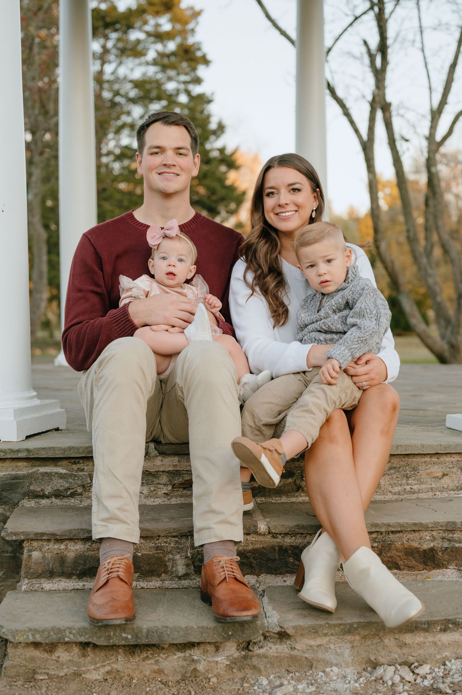 Young family of four with two babies poses on historic stone steps with white columns in autumn.