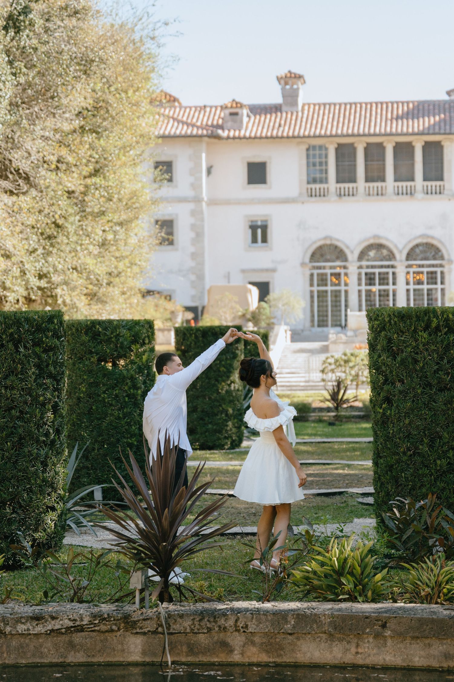 Couple dancing in elegant garden with man spinning woman in white dress near grand historic mansion.