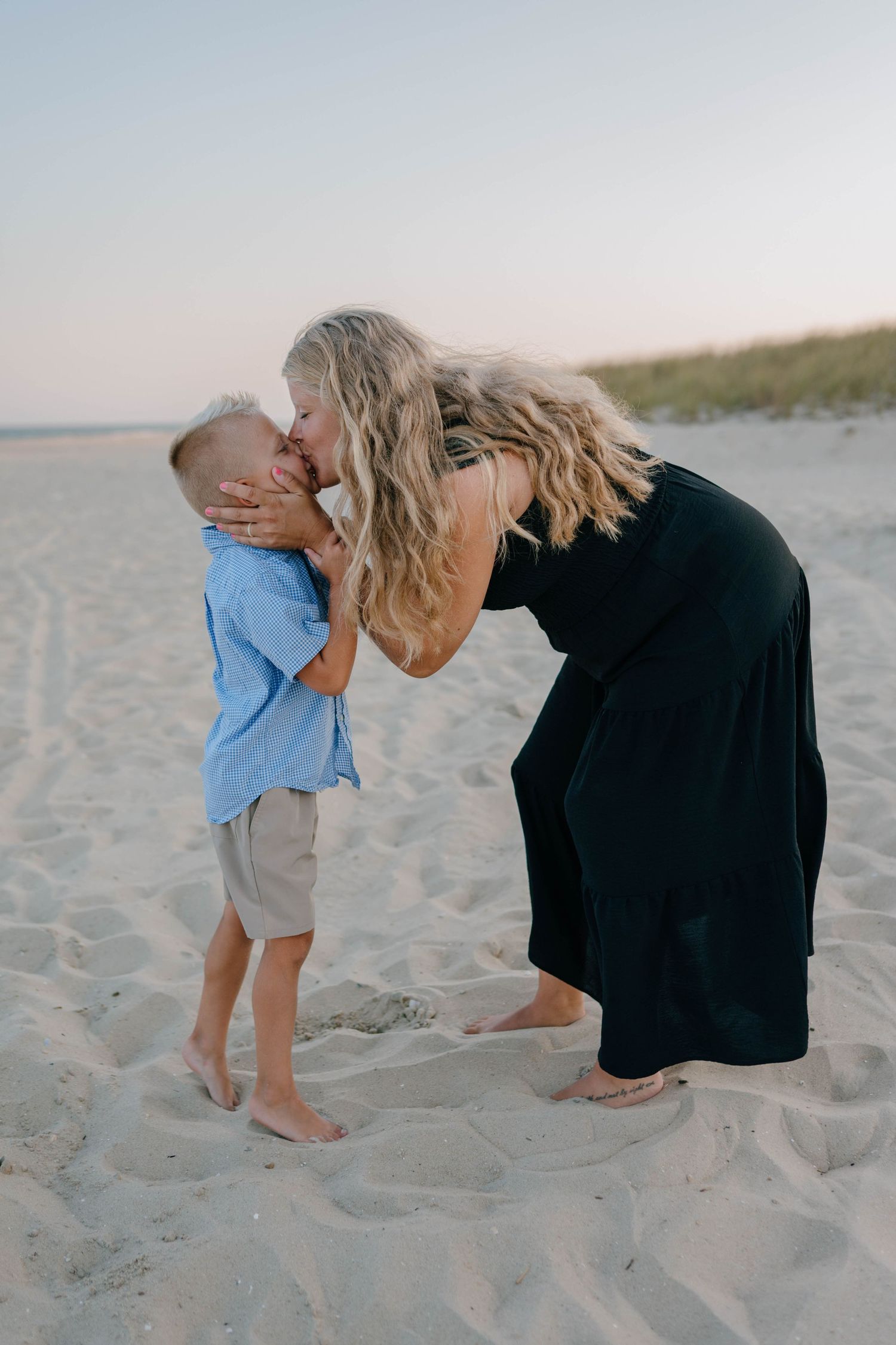 Tender moment as pregnant mother in black dress kisses young blonde son on sandy beach at dusk.