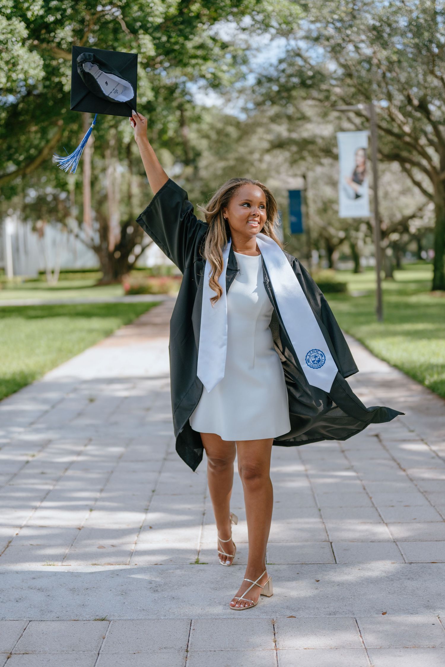 Smiling graduate in black gown raising cap while wearing Lynn University stole on campus path.