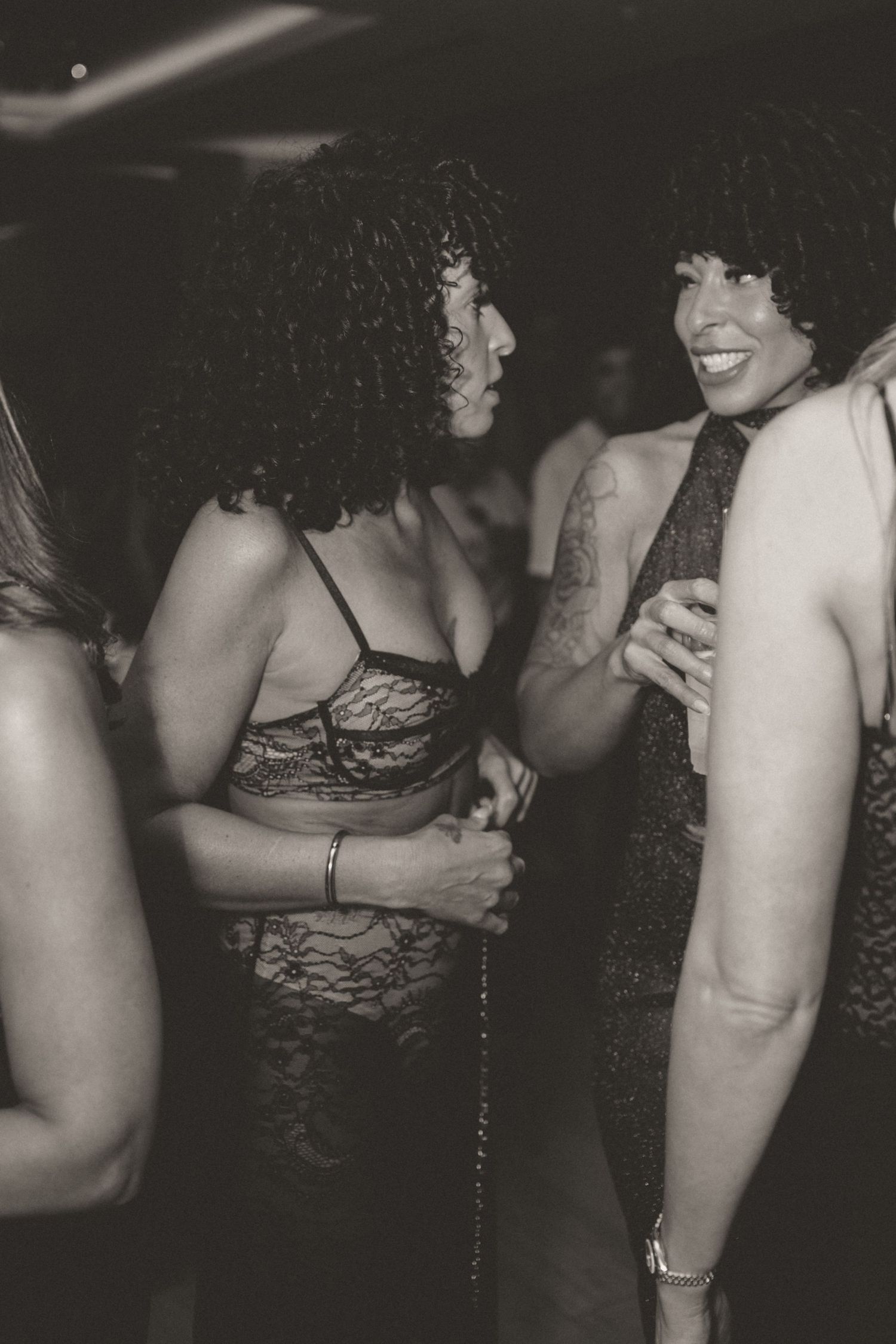 Two women with curly hair chat and laugh together at a nightclub party in black and white photo.