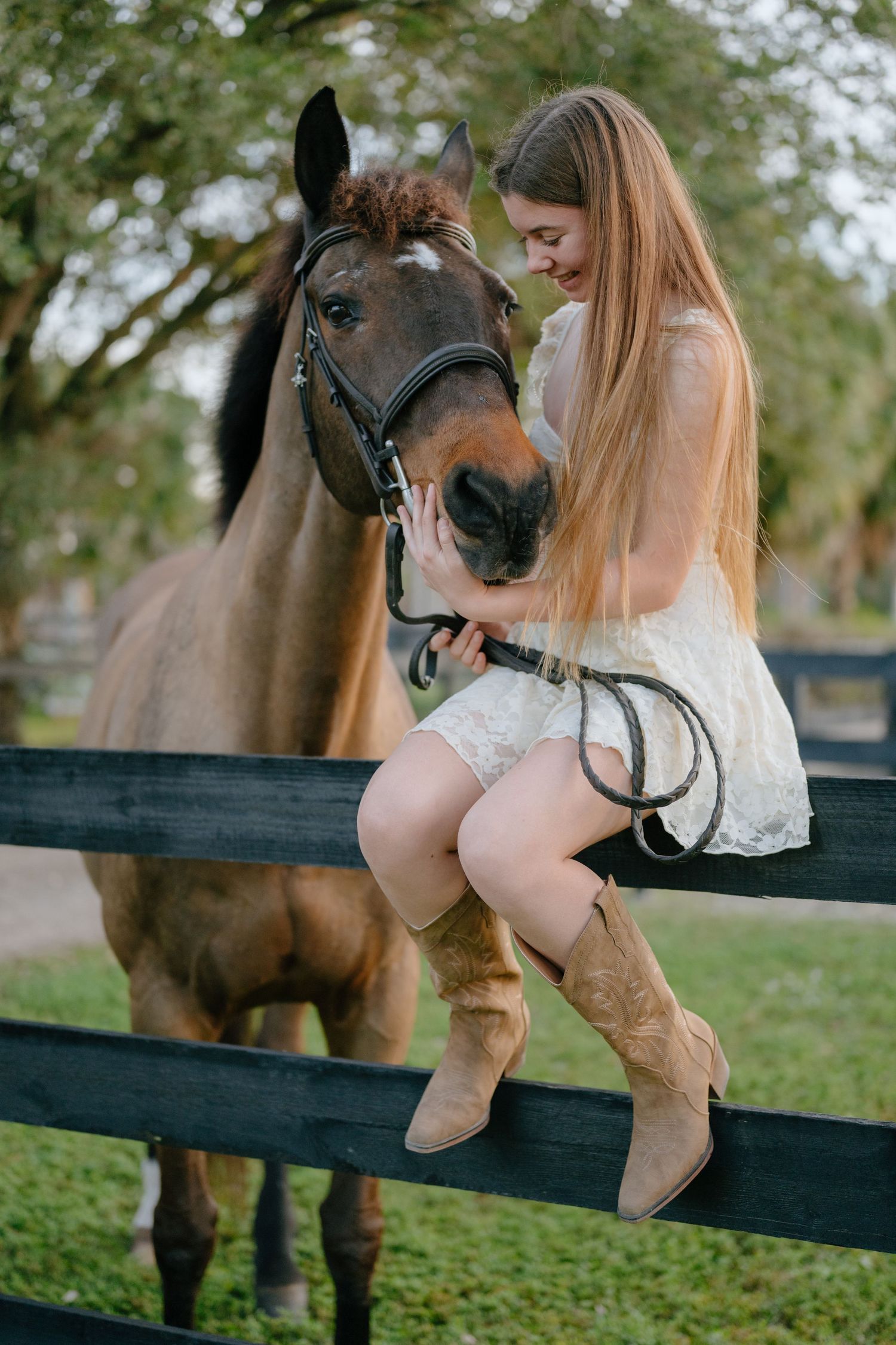 Young woman in white dress and cowboy boots sits on fence lovingly embracing a bay horse.
