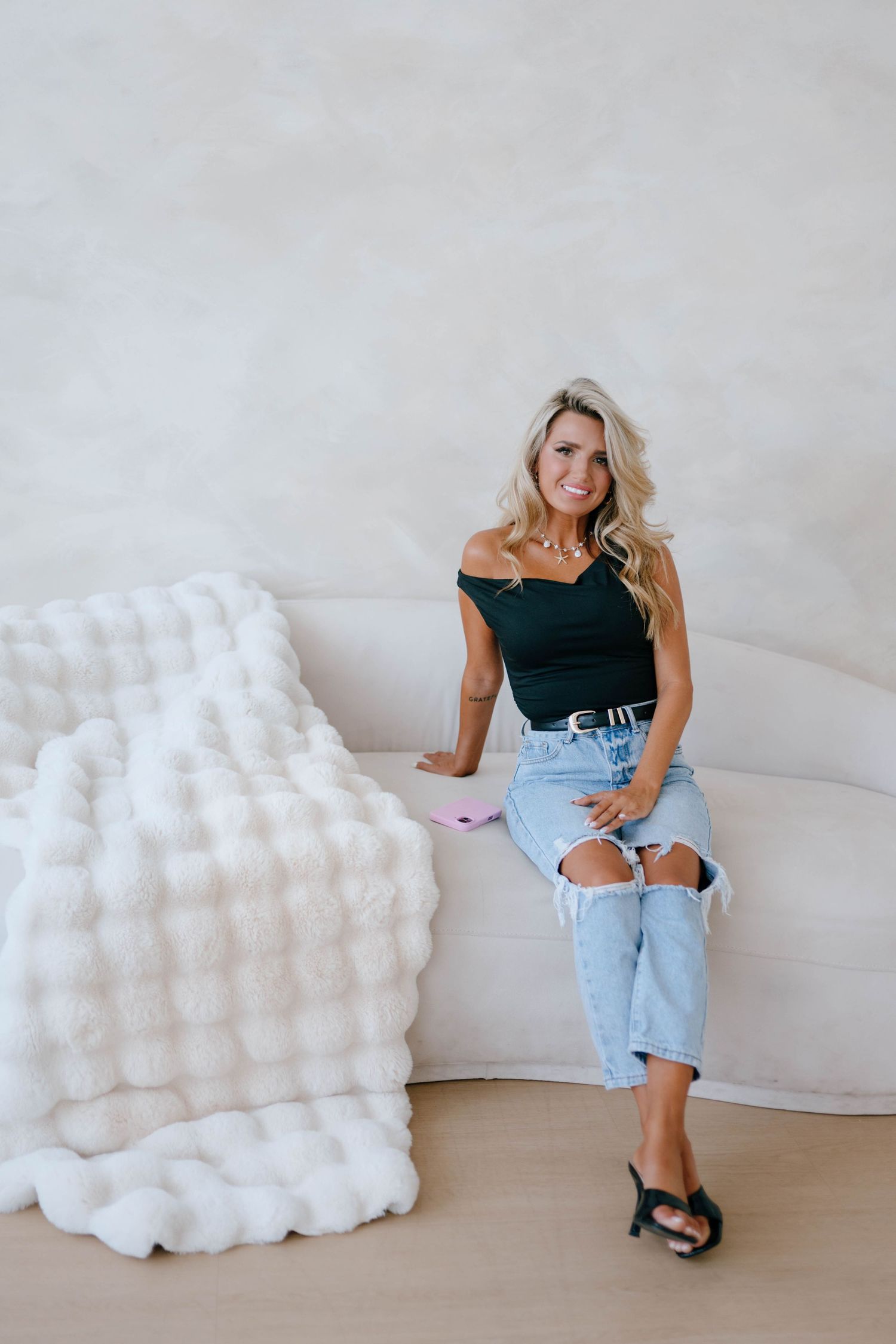 Blonde woman in black off-shoulder top and ripped jeans sitting on white sofa.