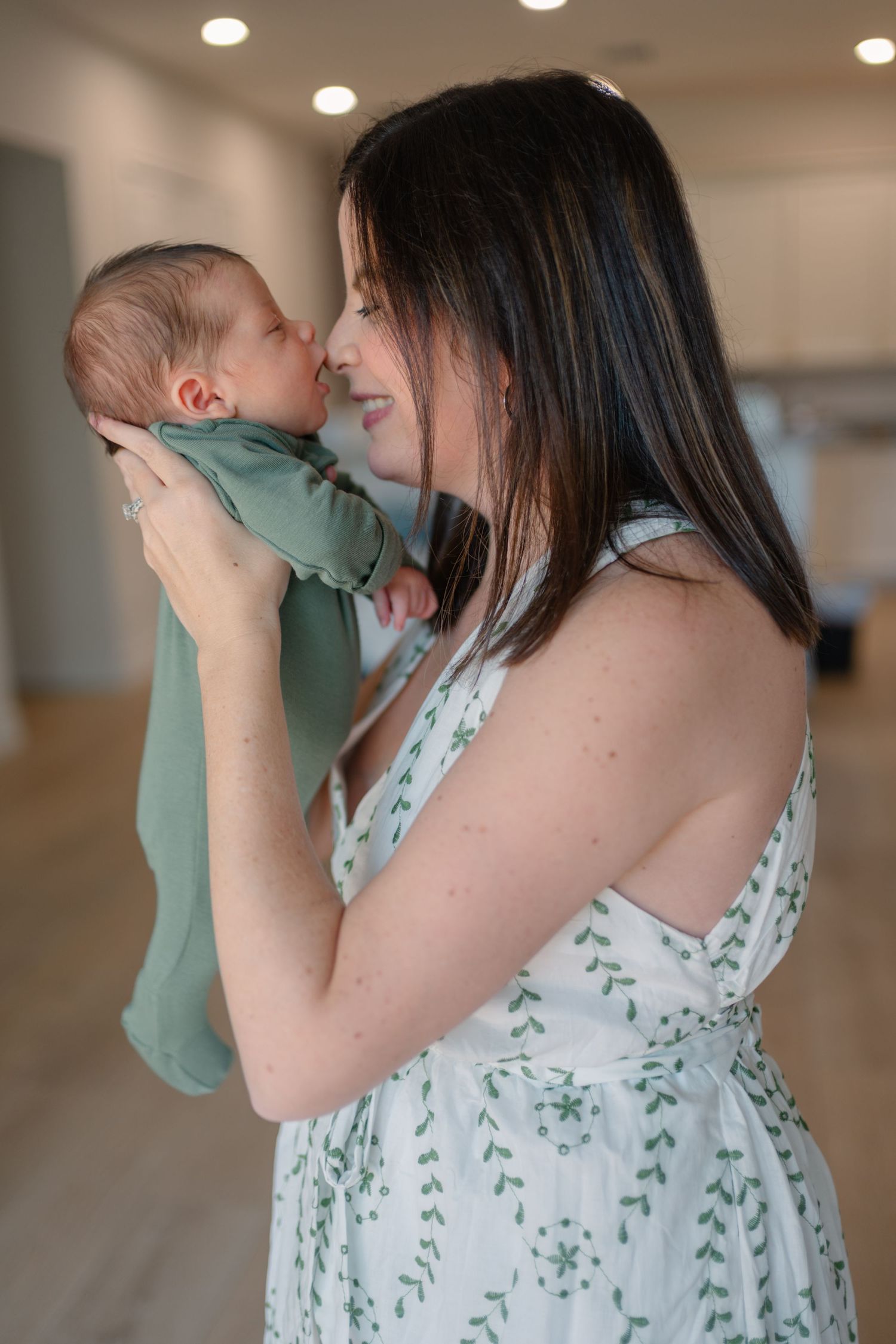 Mother in floral dress gently kisses her newborn baby wrapped in green cloth.