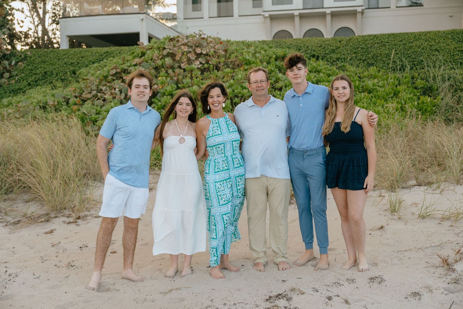 Family of six smiling together on a sandy beach, dressed in coordinating blue and white outfits during golden hour.