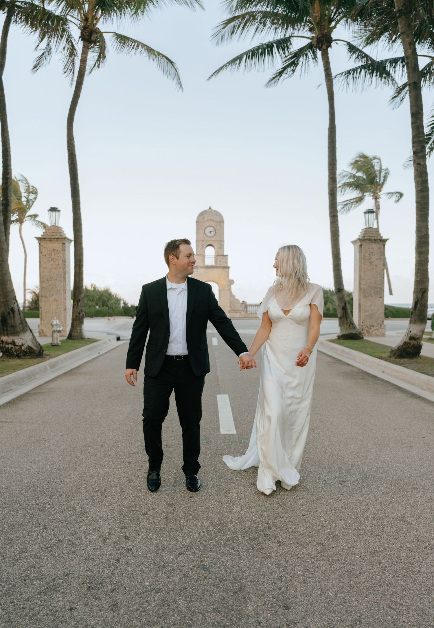 Bride and groom walk hand in hand on a palm-lined road with a historic clock tower backdrop.