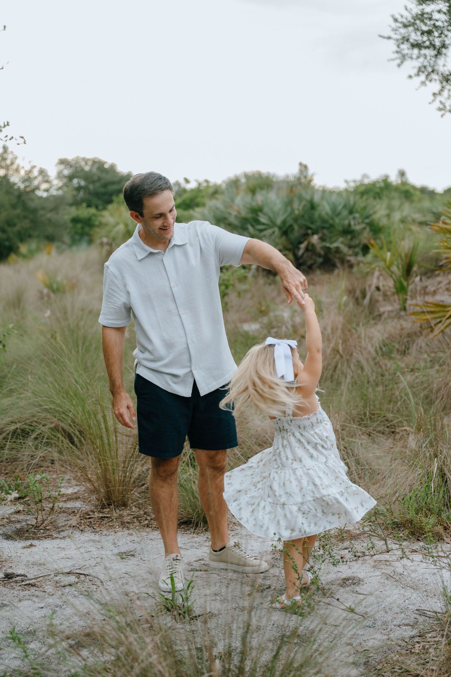 A father spins his young blonde daughter in a floral dress outdoors among tall grasses.