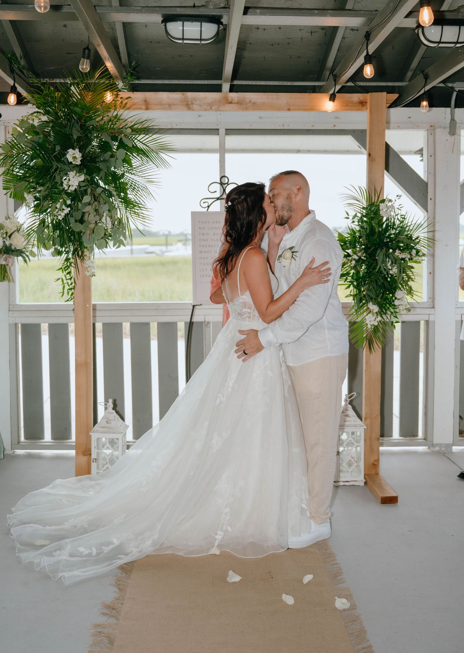 Bride and groom share first kiss under wooden arch decorated with tropical greenery at beach wedding.