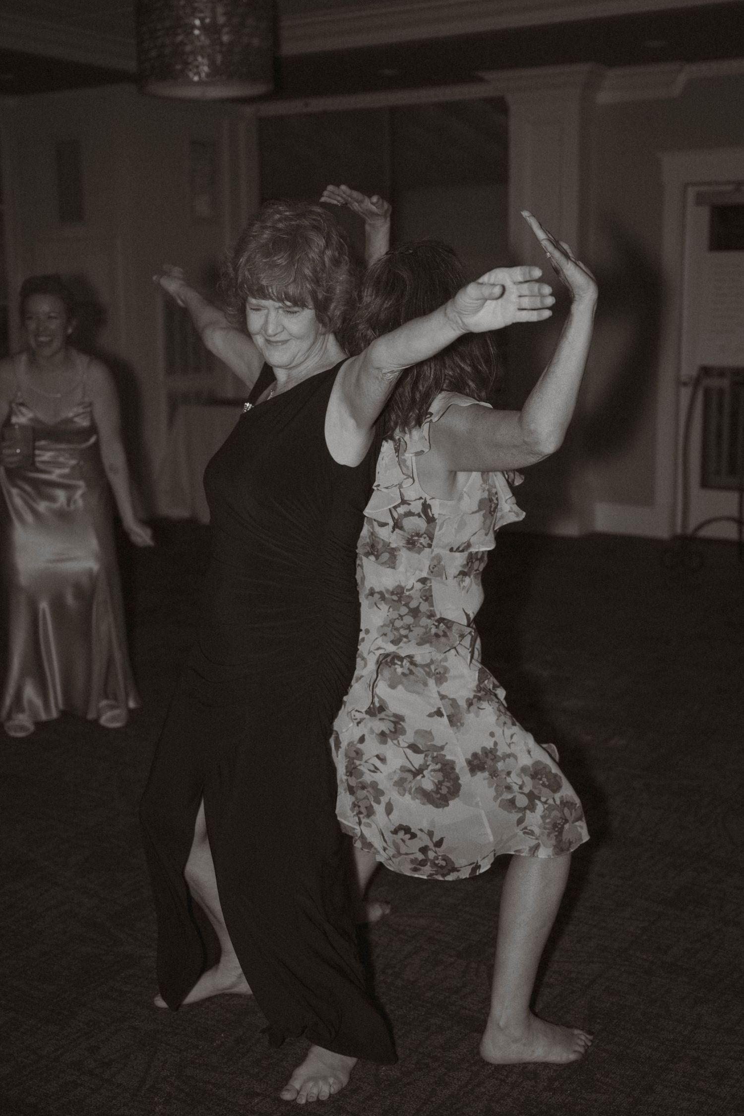 Black and white photo of two women dancing barefoot together at lively wedding reception celebration.