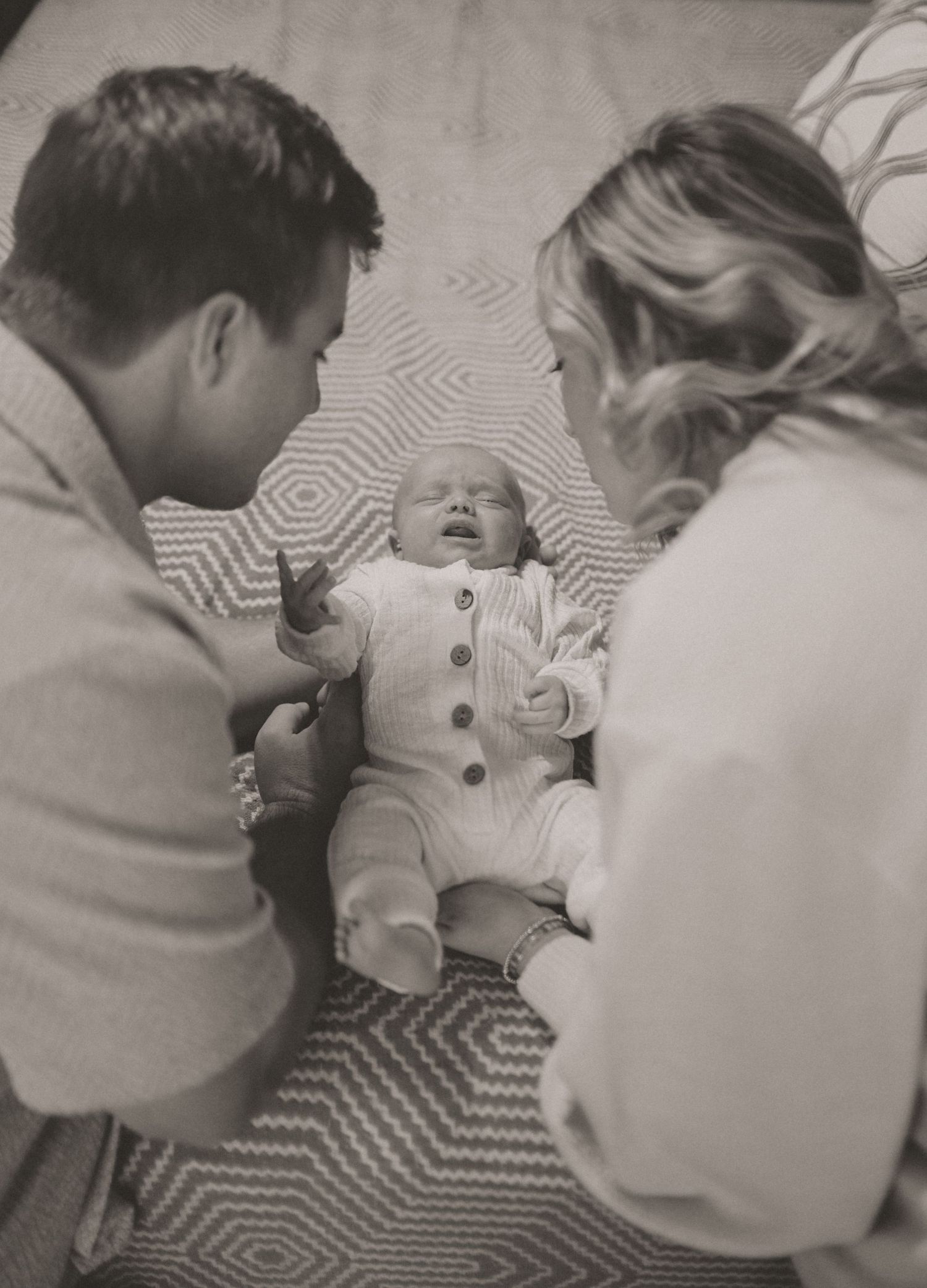 Black and white overhead photo of two parents looking down at their newborn baby on a blanket.