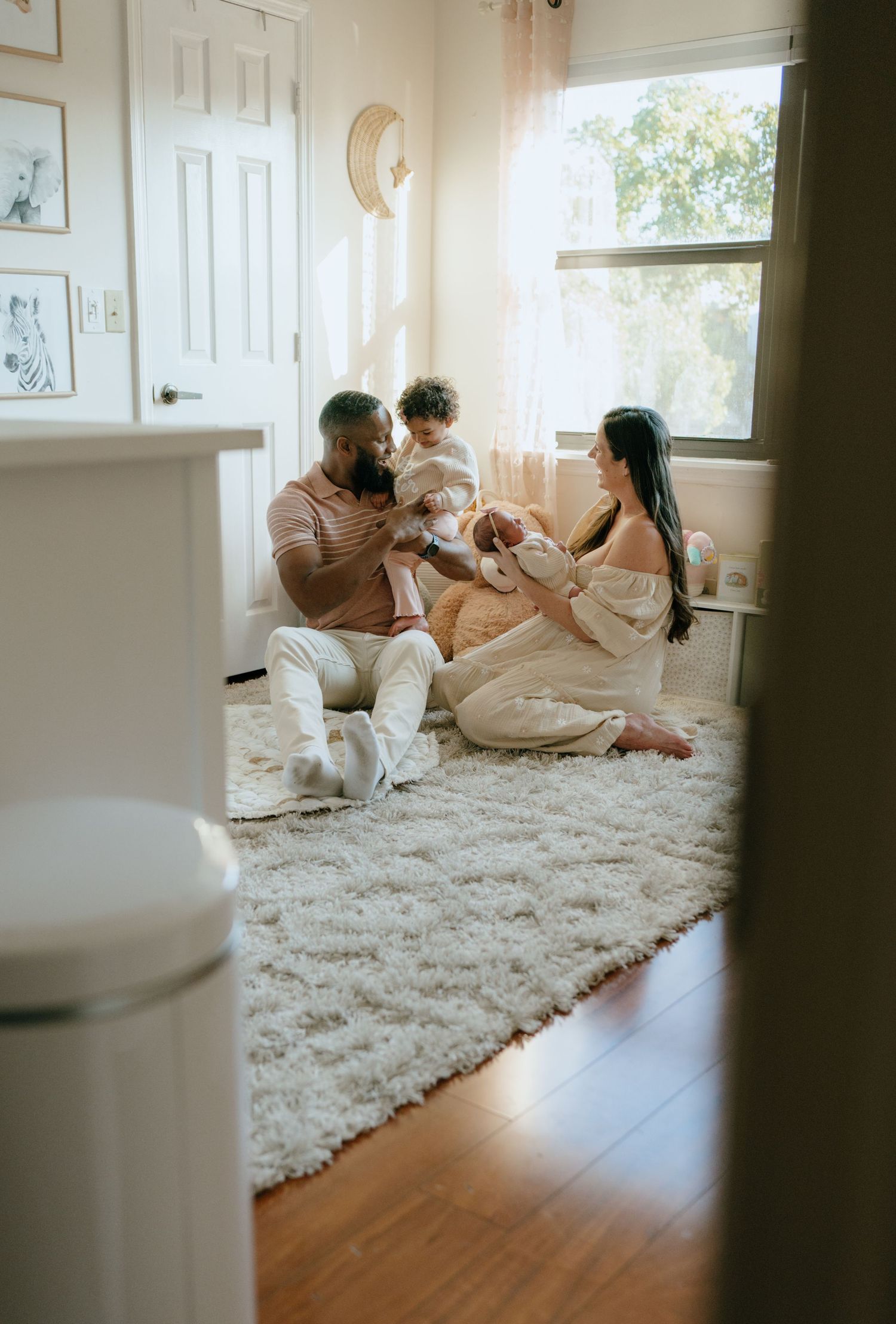 Family of four sitting together on a nursery floor in warm natural light.