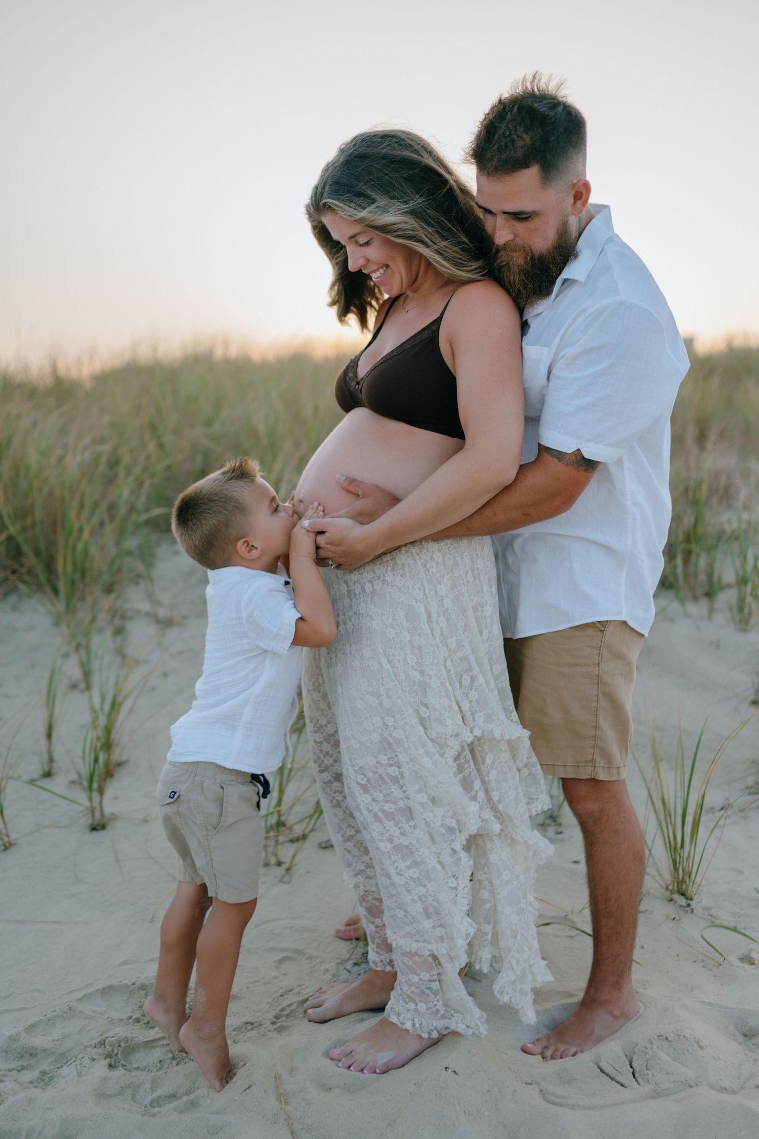 A happy family maternity photoshoot on sandy beach dunes at golden hour sunset.