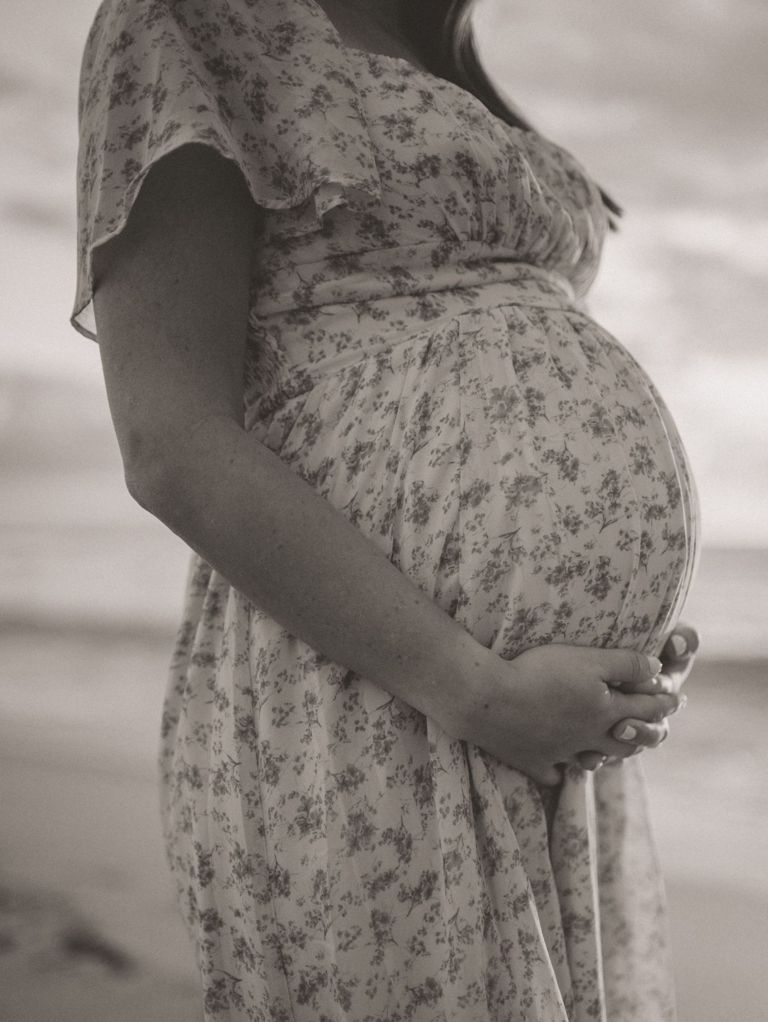 Black and white close-up of pregnant woman in floral dress holding baby bump at beach.