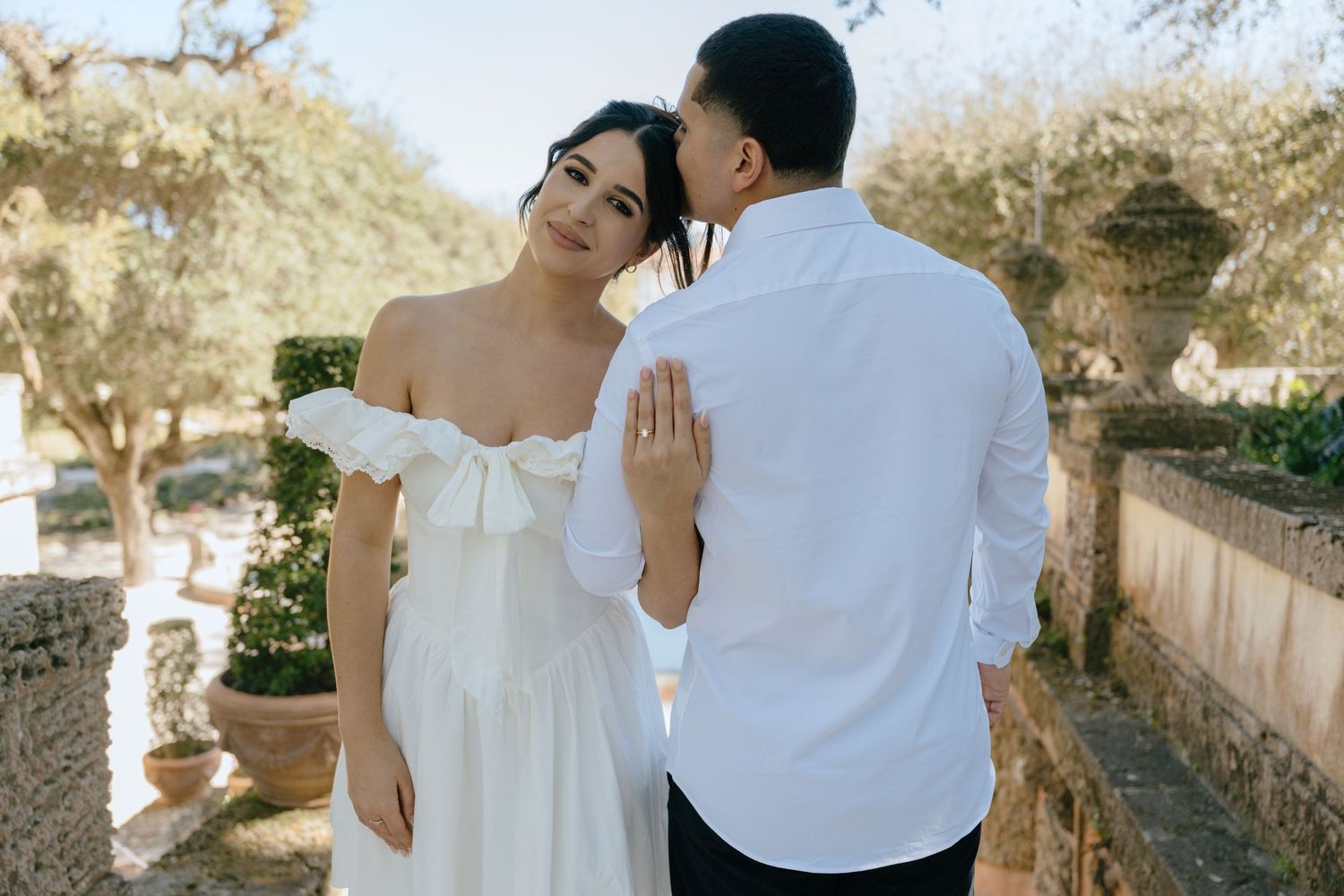 Couple in white outfits pose romantically in a garden, bride in off-shoulder gown looks back at groom.