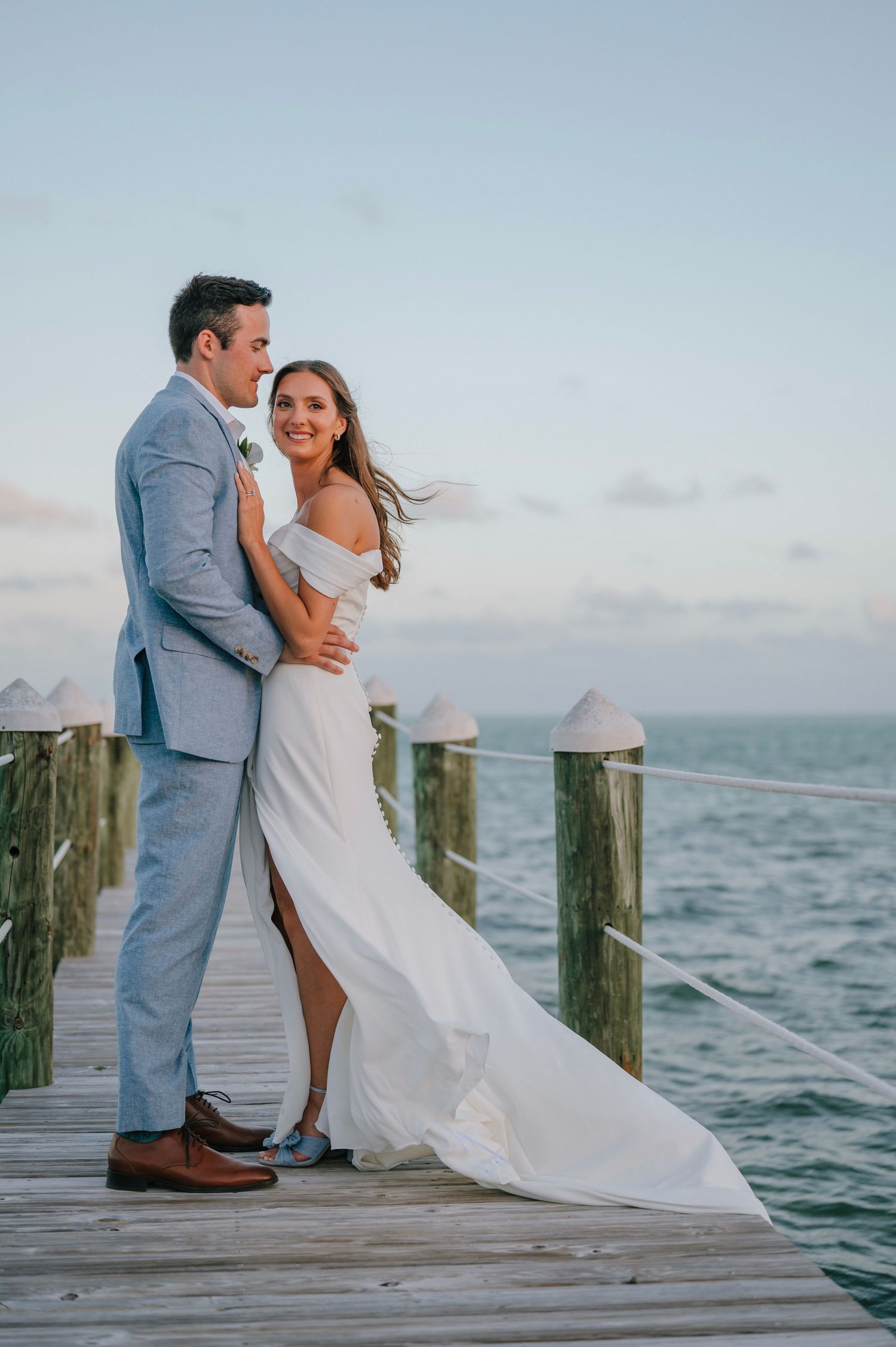 Bride and groom embracing on a wooden dock at sunset, bride wearing an off-shoulder gown by the ocean.