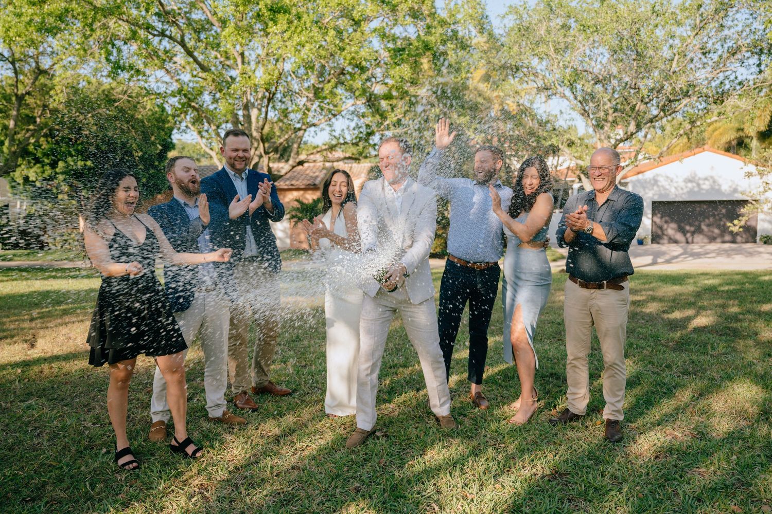 Wedding party celebrates with champagne spray outdoors in a sunny garden with joyful guests.