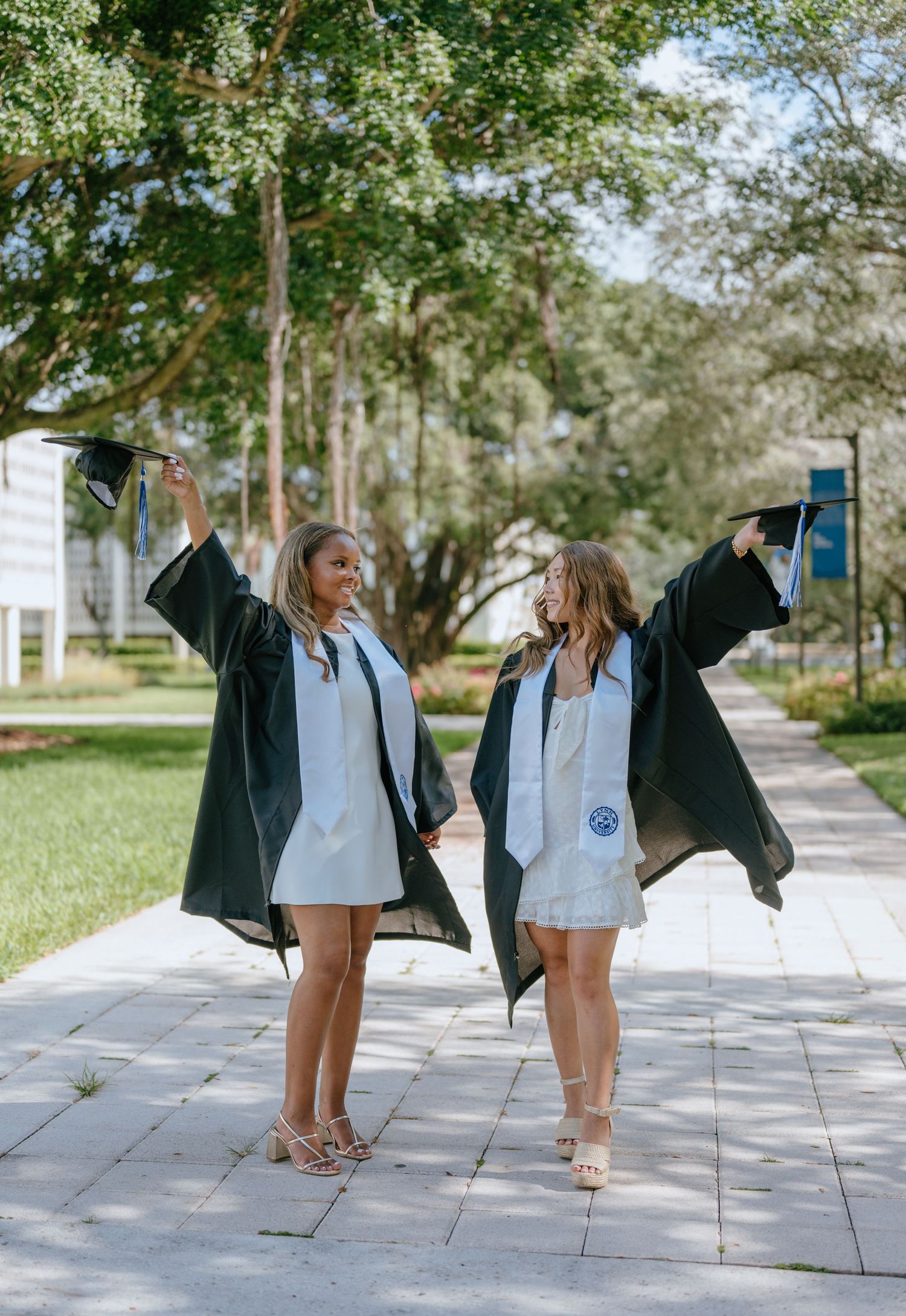Two smiling graduates in black gowns raising caps triumphantly on a sunlit university campus path.