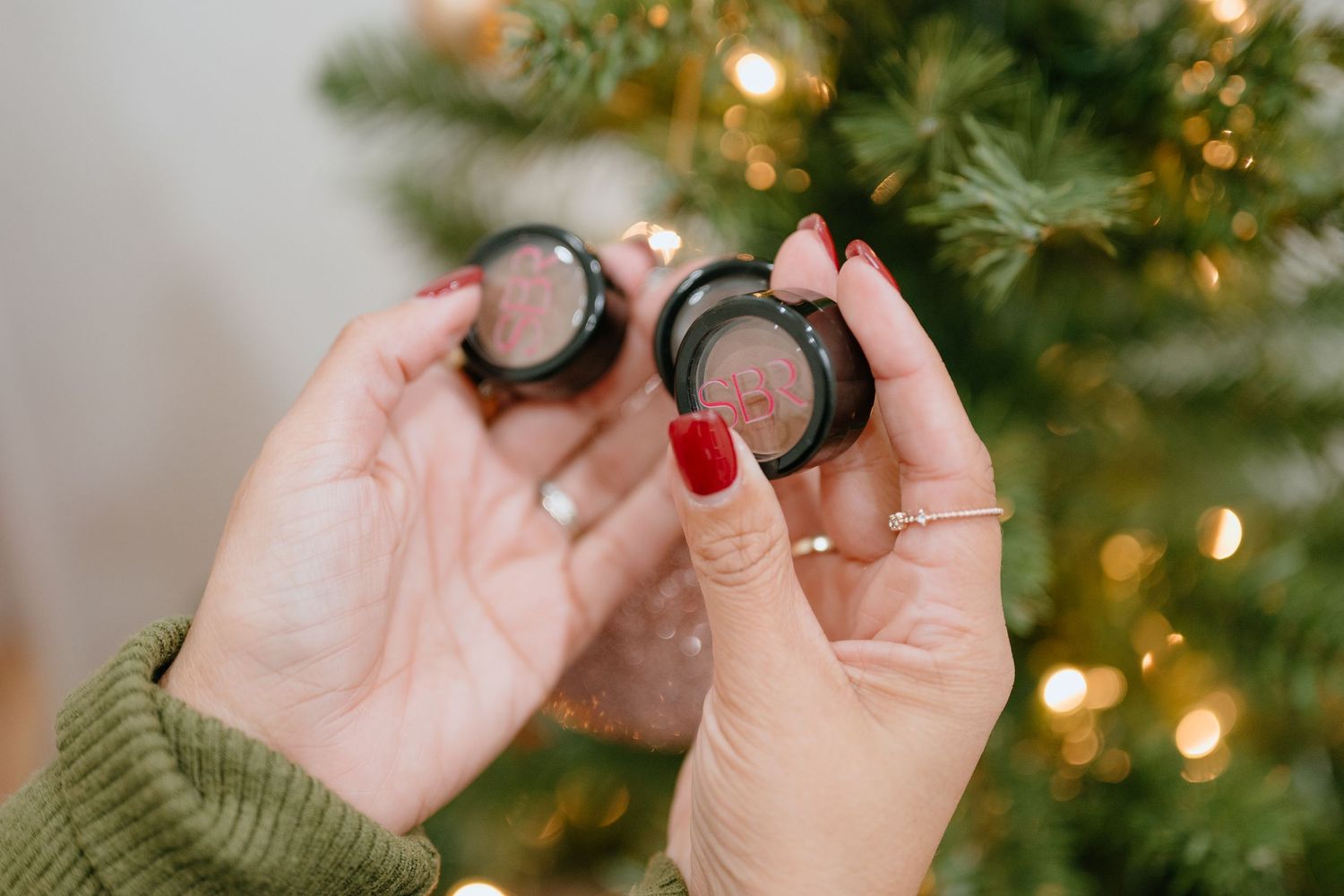 Hands in green sweater holding two black compact brow or eyeshadow products near a lit Christmas tree.