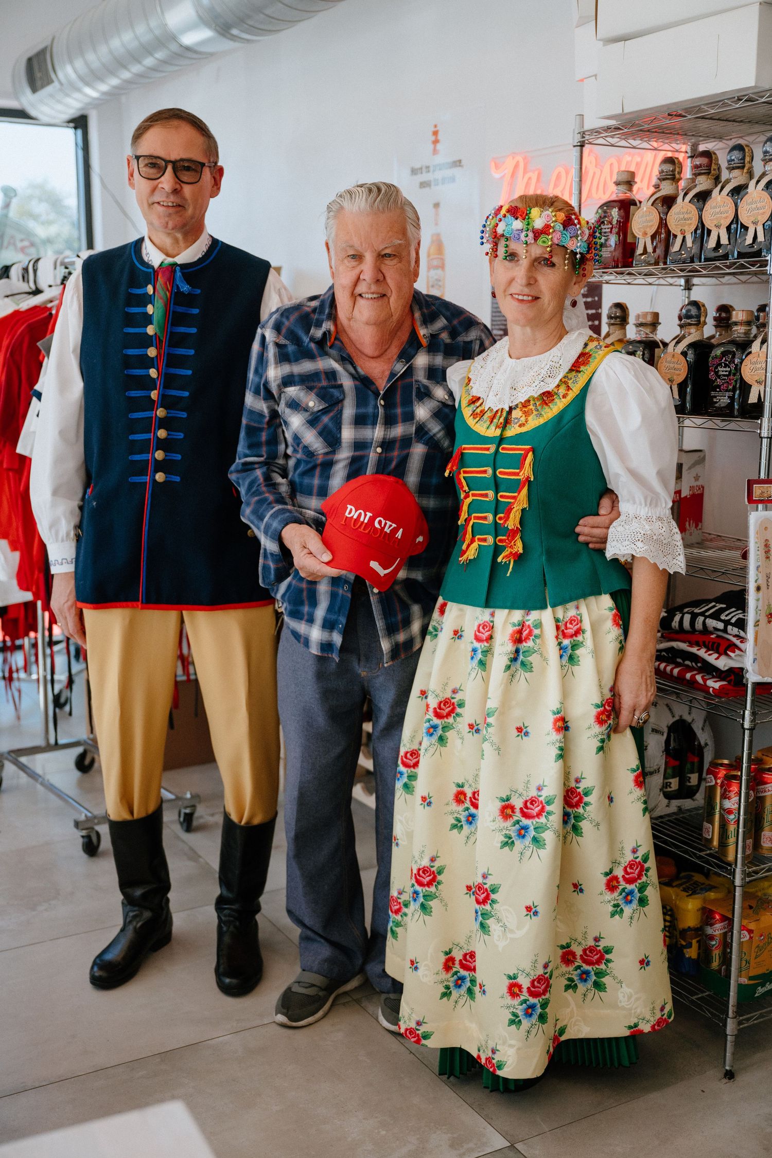 Three people posing in traditional Polish folk costumes inside a Polish grocery store.