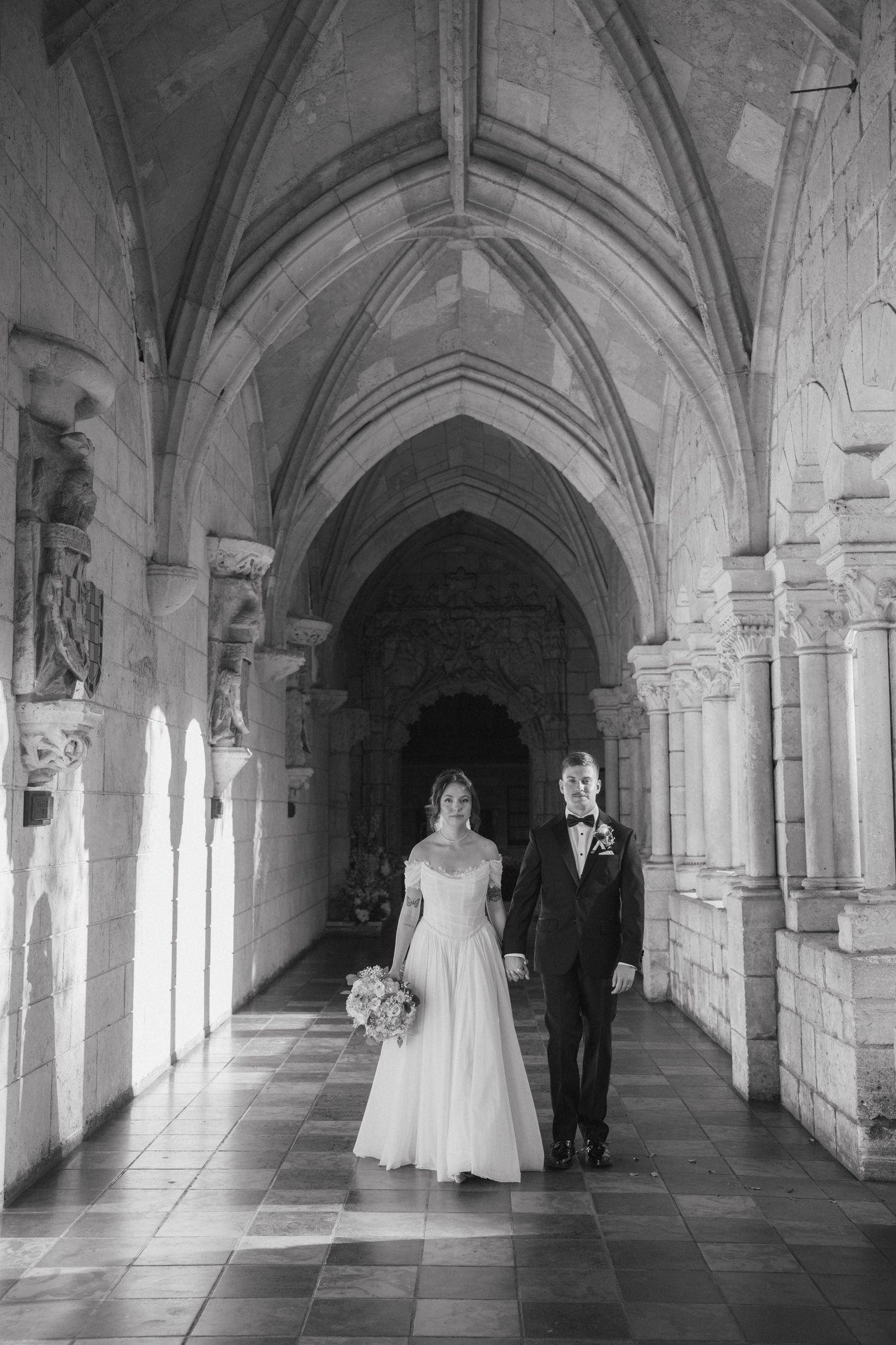 Black and white photo of bride and groom walking through vaulted Gothic stone cathedral hallway.