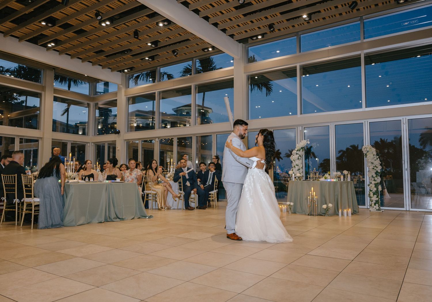 Bride and groom share their first dance in a stunning glass-walled ballroom at evening reception.