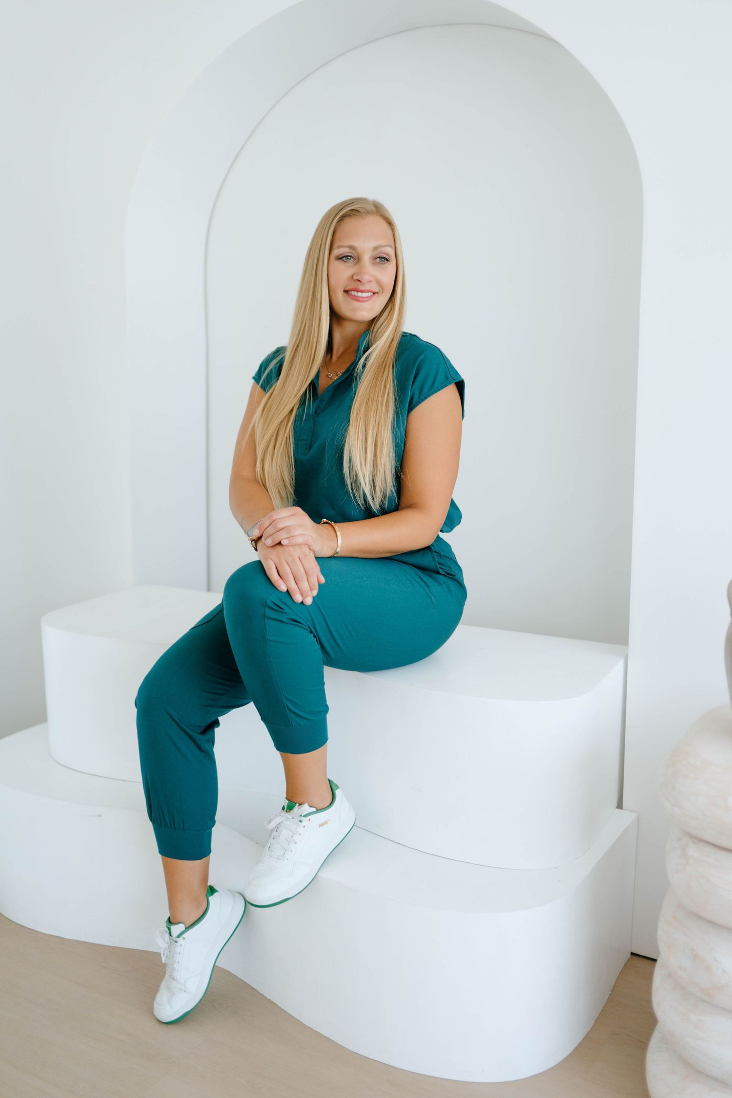 Smiling blonde woman in teal scrubs sitting on a white architectural bench inside a curved arch alcove.