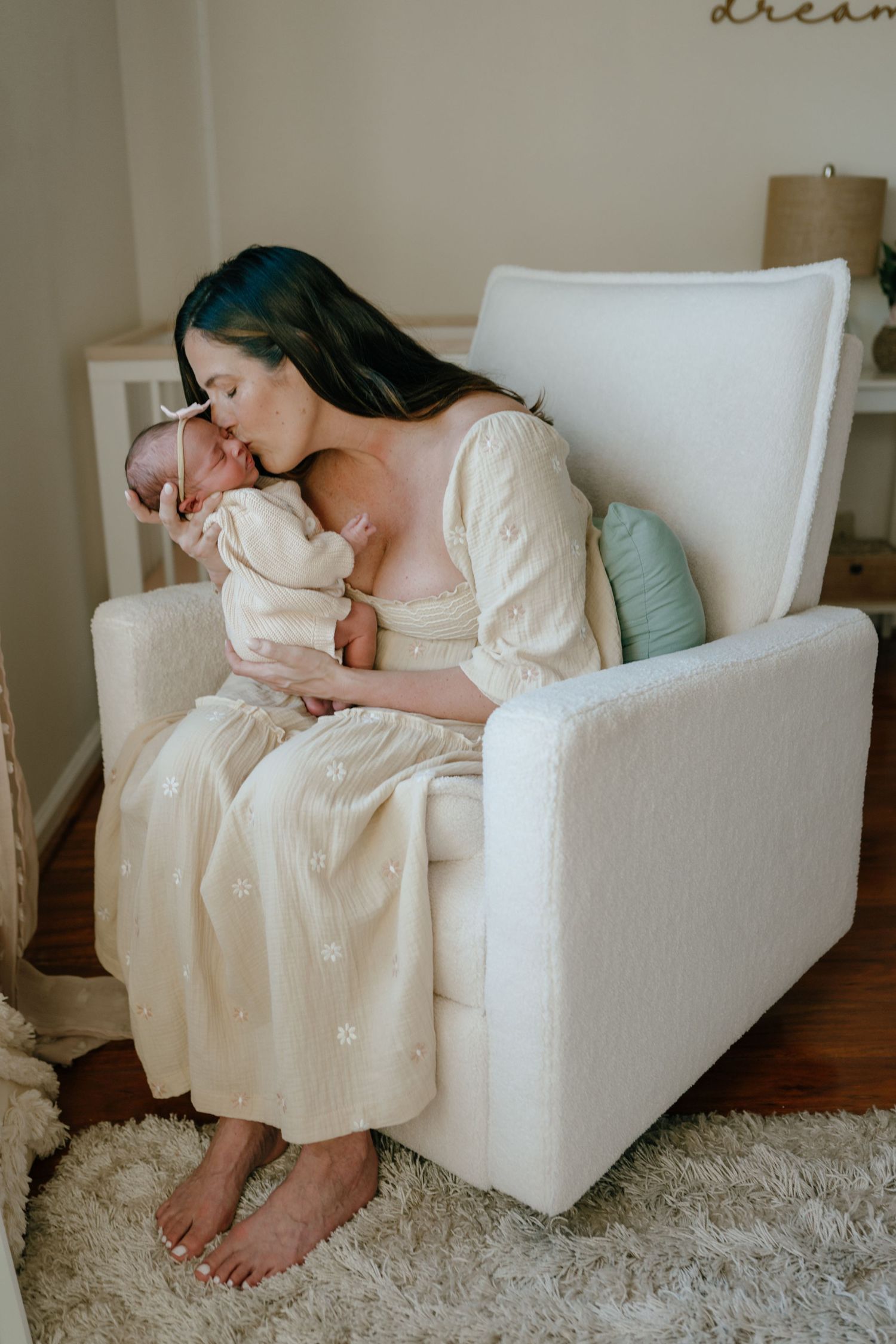 Mother in cream dress sitting in nursery glider chair kissing sleeping newborn baby.