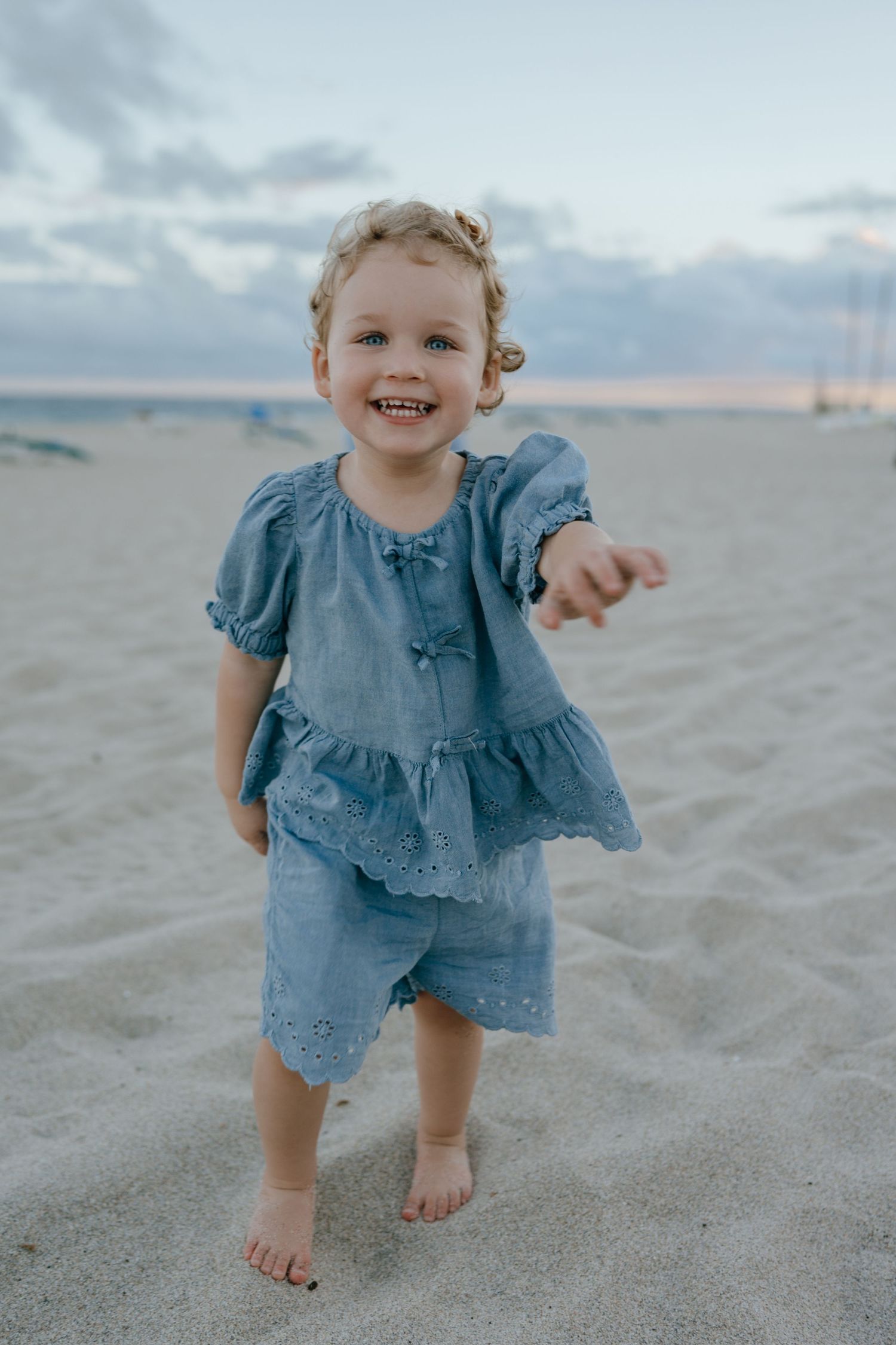 Adorable curly-haired toddler girl in blue outfit laughing and walking barefoot on sandy beach at sunset.