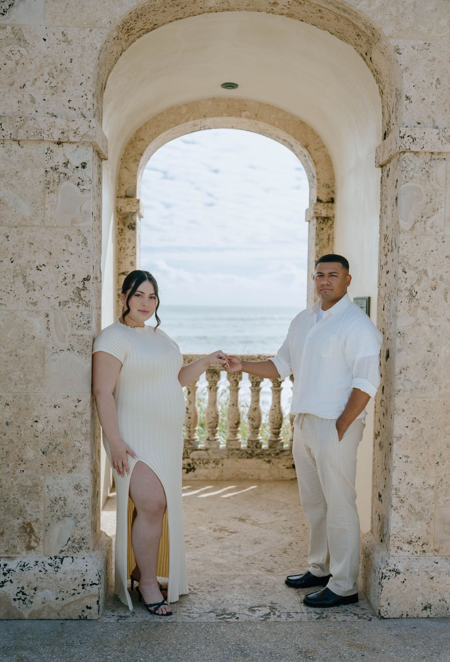 Couple in white attire holding hands under a stone archway with ocean views at a coastal venue.