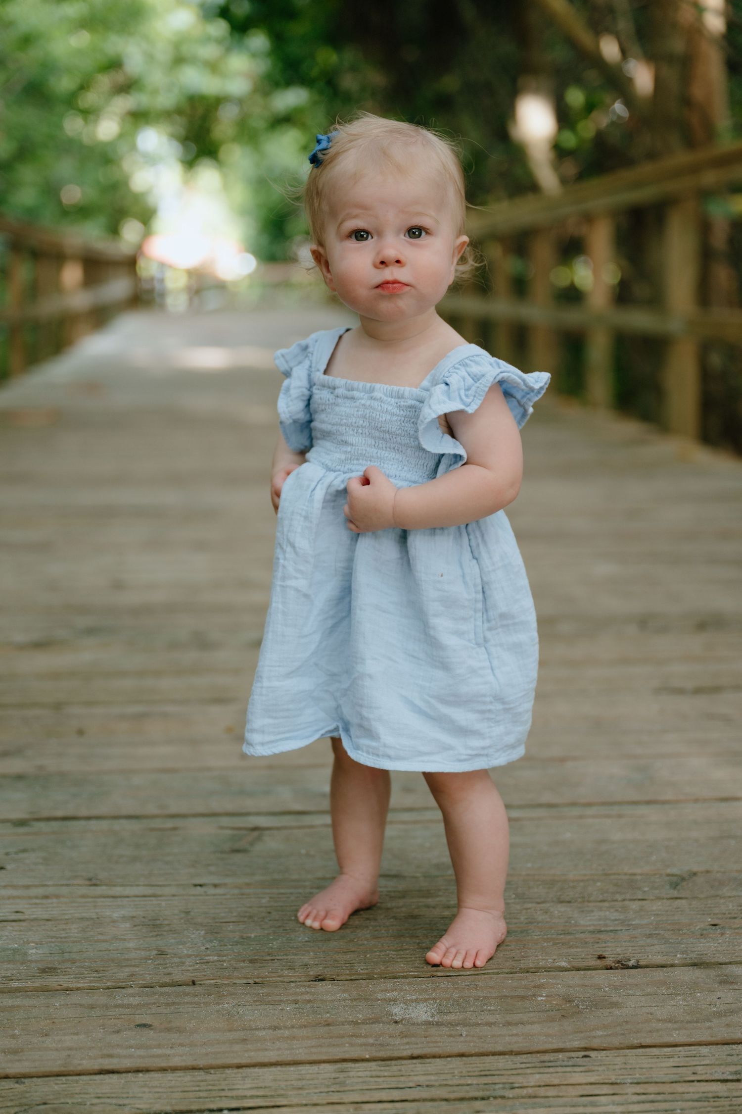 A barefoot toddler girl in a light blue dress stands alone on a wooden boardwalk outdoors.