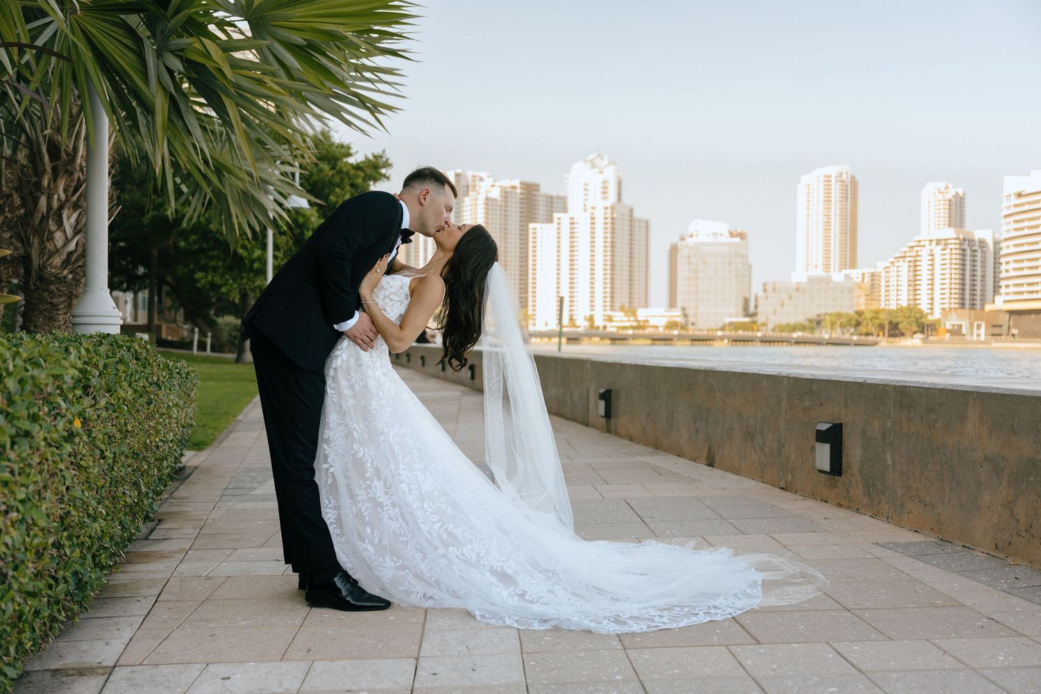 Bride and groom sharing a romantic dip kiss along a waterfront promenade with city skyline.