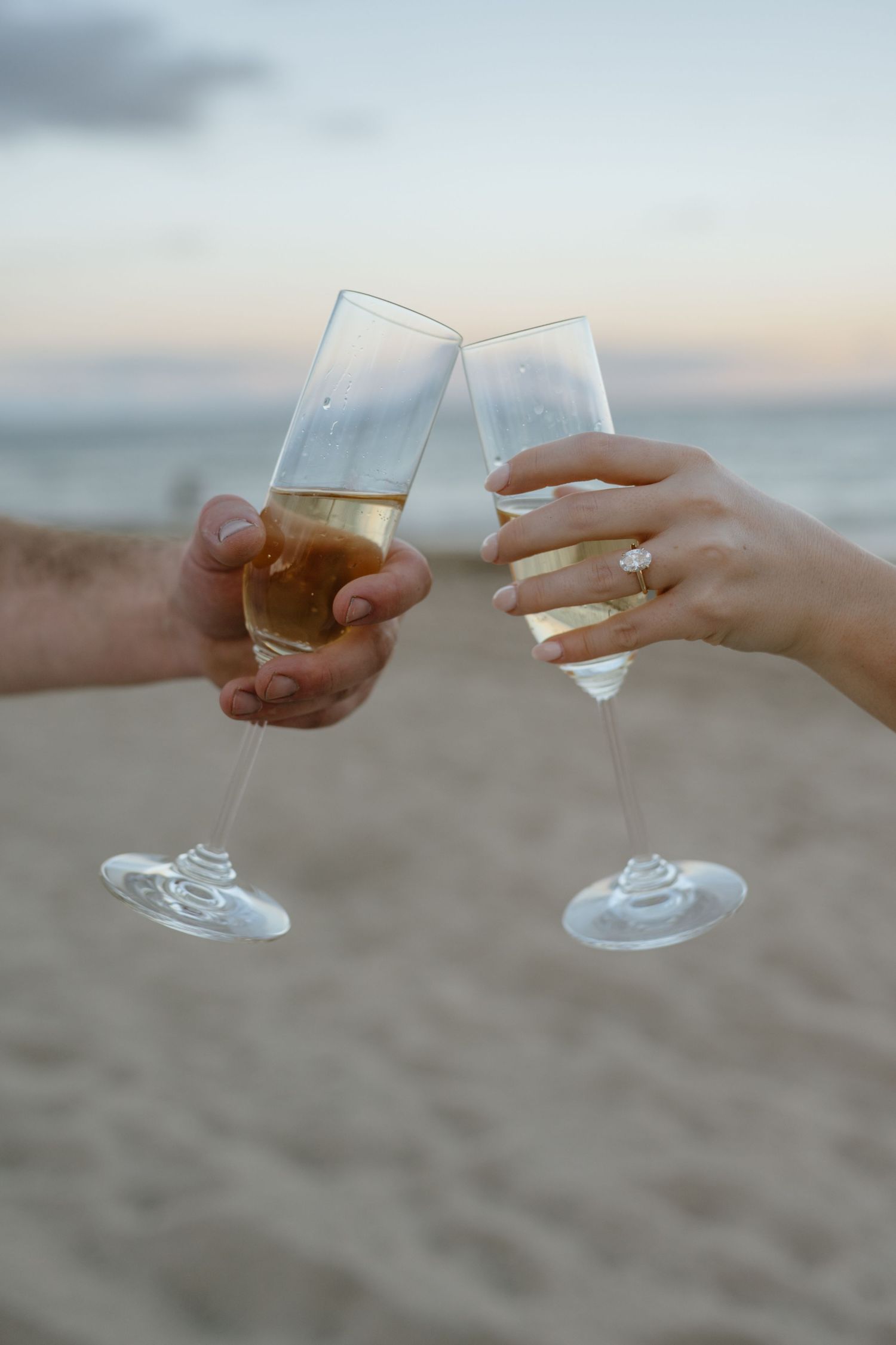 Close-up of an engaged couple clinking champagne flutes on the beach, showing off an engagement ring.
