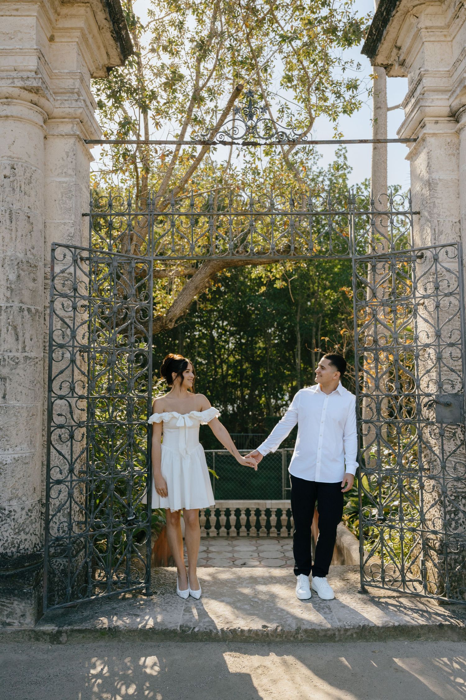 Engaged couple holding hands framed by ornate iron gates at a historic estate garden entrance.