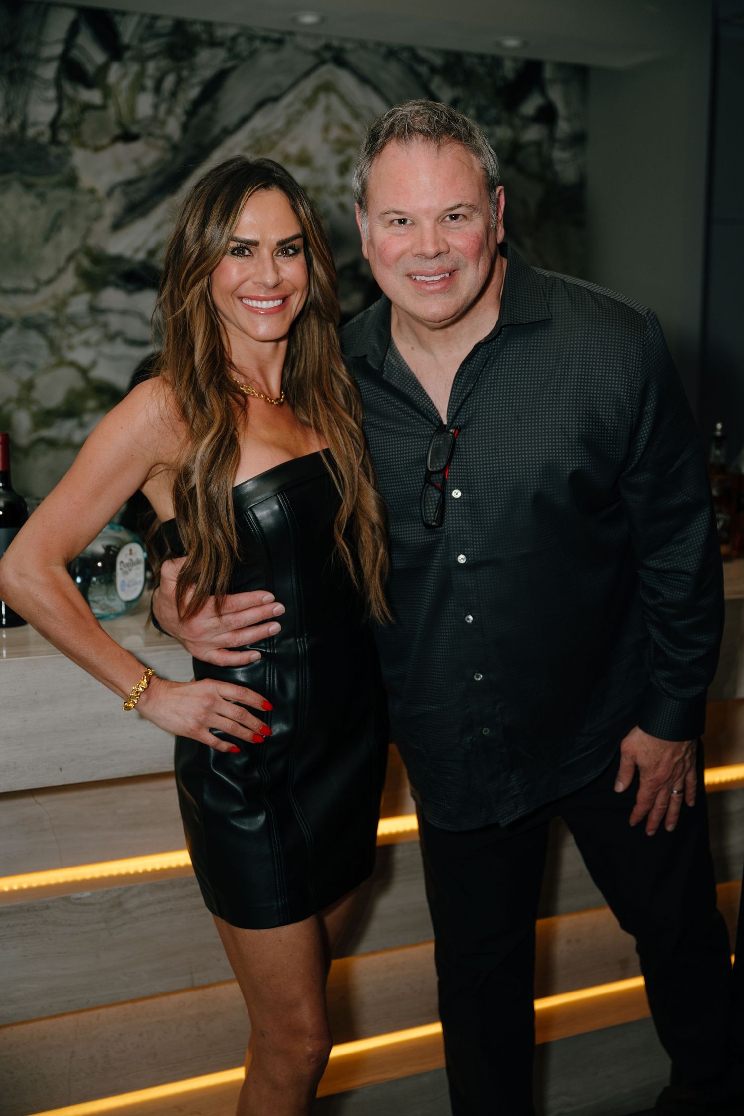 Smiling couple poses together at an upscale indoor event, woman in black leather dress and man in black shirt.